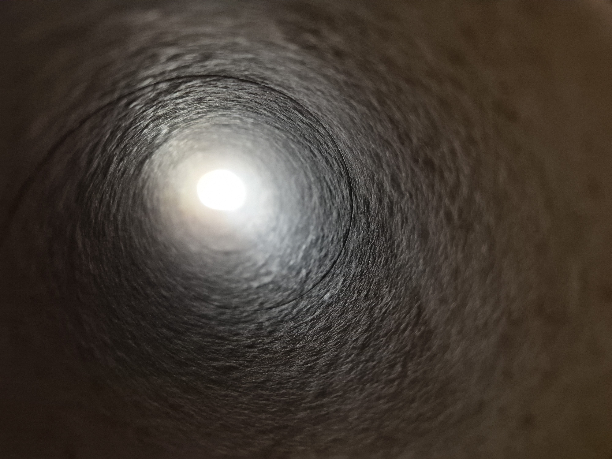 The image depicts a close-up, low-angle shot looking down the interior of a cylindrical object, possibly a cardboard tube. The walls of the tube are textured and appear to be a dark, muted brown color, with visible striations and variations in shading. Light is emanating from the far end of the tube, creating a bright, circular focal point that diminishes in intensity as it recedes into the darkness. The overall impression is one of depth and perspective, drawing the viewer's eye towards the distant light source. There are no people or specific landmarks visible in this image. The scene is entirely abstract and focuses on the texture and form of the interior of the tube and the light at its end.