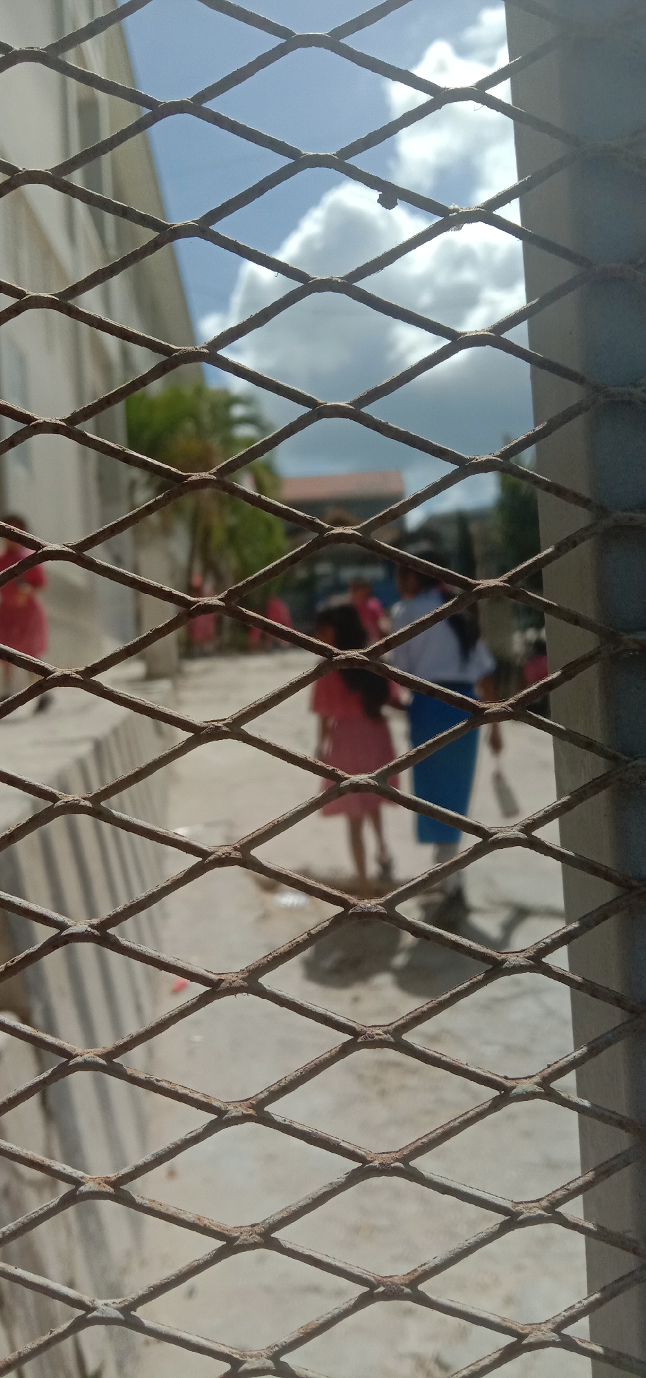 The image, taken through a rusted metal grate, depicts a daytime scene on what appears to be a school campus in Kendari, Indonesia. The background is softly out of focus, showing a light-colored building on the left and other structures and trees further back. Several individuals, primarily children and possibly an adult, are visible walking or standing on a paved pathway. One child in the foreground on the right is wearing a pink dress, and another person to their left is wearing a white shirt and a blue skirt, possibly a school uniform. Shadows are cast on the ground, indicating sunlight. The sky above is partly cloudy. The foreground is dominated by the diamond-shaped pattern of the metal grate, with some rust visible on its surface.