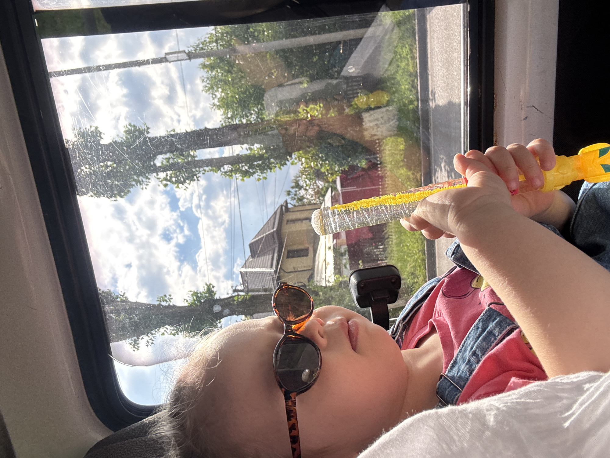 This media file shows a young child lying down inside a vehicle, looking upwards. The child is wearing sunglasses with brown frames, resting on their nose. They are dressed in a pink shirt with a denim overall strap visible. In the child's right hand, they are holding a bubble wand. The bubble wand is yellow and appears to have a loop of bubbles already formed.
The background visible through the vehicle's window shows a bright, sunny sky with fluffy white clouds. Trees and a building are reflected on the glass, suggesting an outdoor scene. The setting appears to be daytime, likely mid-morning or afternoon, due to the bright sunlight. The reflection on the window suggests the vehicle is in motion or parked near trees and a residential area. There is no visible text in the media file. The overall mood is relaxed and playful, with the child engaged with the bubble wand. The location is Mikhaylovsk, Russia.