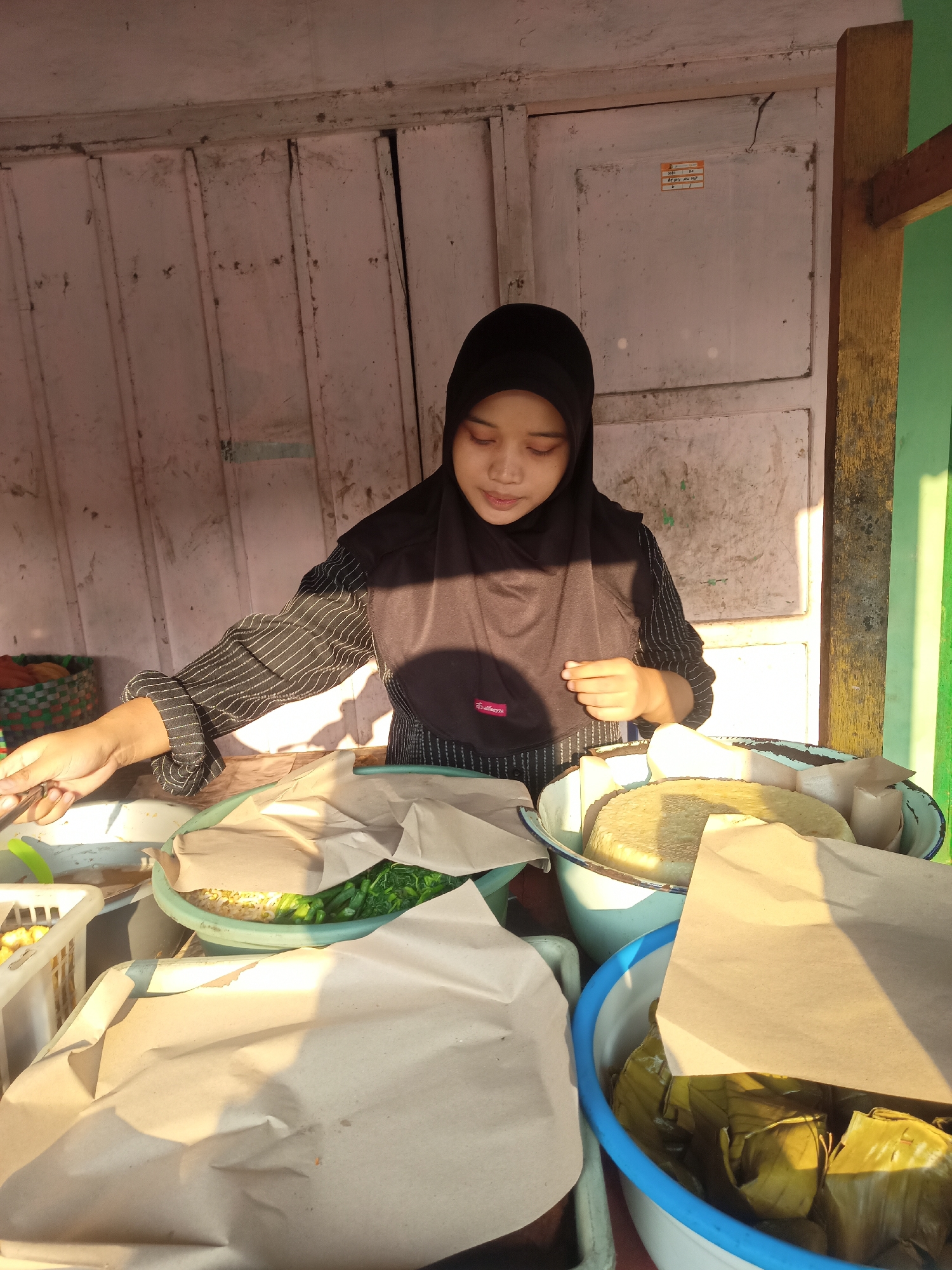 In Gemolong, Indonesia, a young woman wearing a black hijab and a striped shirt stands behind a stall. She is preparing food, with her right hand holding a metal utensil and her left hand poised over a selection of dishes. The stall displays various food items, including what appears to be cooked rice, green vegetables, and wrapped parcels. These are arranged in bowls and containers, some covered with brown paper. The stall itself is constructed with light-colored wooden panels, showing signs of wear and tear. The overall scene suggests an outdoor food vendor operating in a local marketplace or street setting.