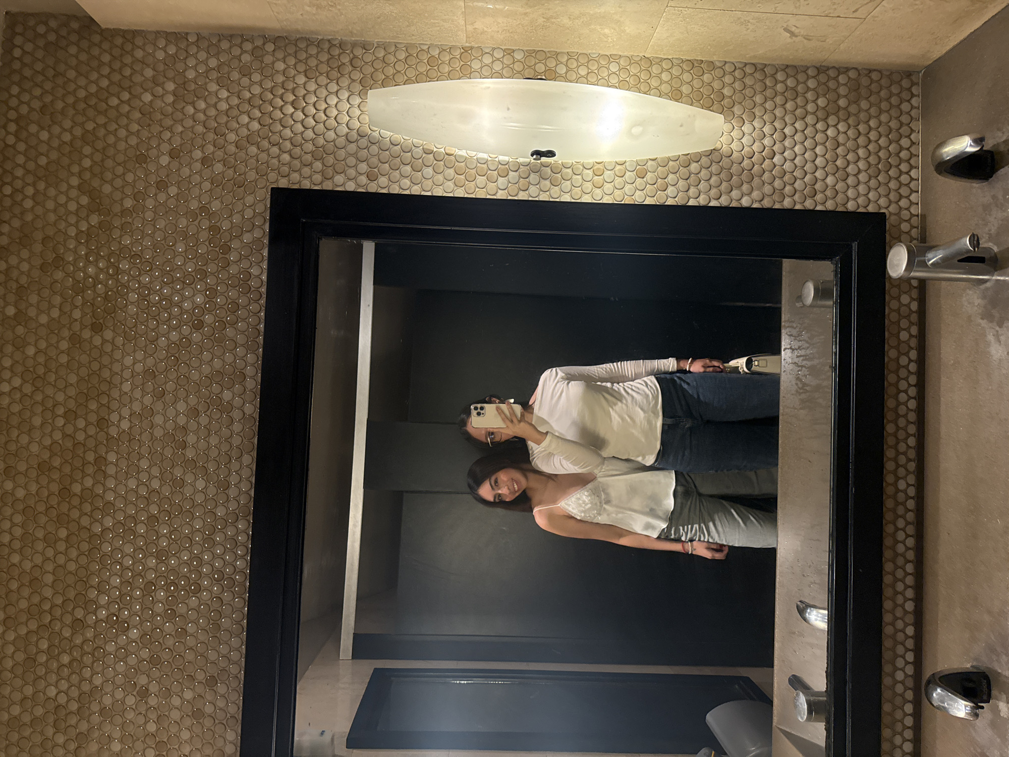 Here's a detailed description of the image:
**Setting:** The photograph appears to have been taken in a bathroom. The walls are tiled with small, circular, light-brown tiles. A long, oval light fixture is positioned above a mirror.
**Mirror and Reflection:** The image shows a large mirror with a black frame. Two women are reflected in the mirror, appearing to be taking a selfie.
**People:**
* **Woman 1:** She is wearing a white long-sleeved shirt and blue jeans. She is holding a cell phone, presumably taking the picture.
* **Woman 2:** She is wearing a white tank top and light-colored pants. She is smiling at the camera.
**Objects:**
* Mirror
* Cell phone
* Light fixture
* Towel holder
* Sink
* Toilet
**Location in Mexico City:** There is not enough information in the image to determine the exact location in Mexico City.