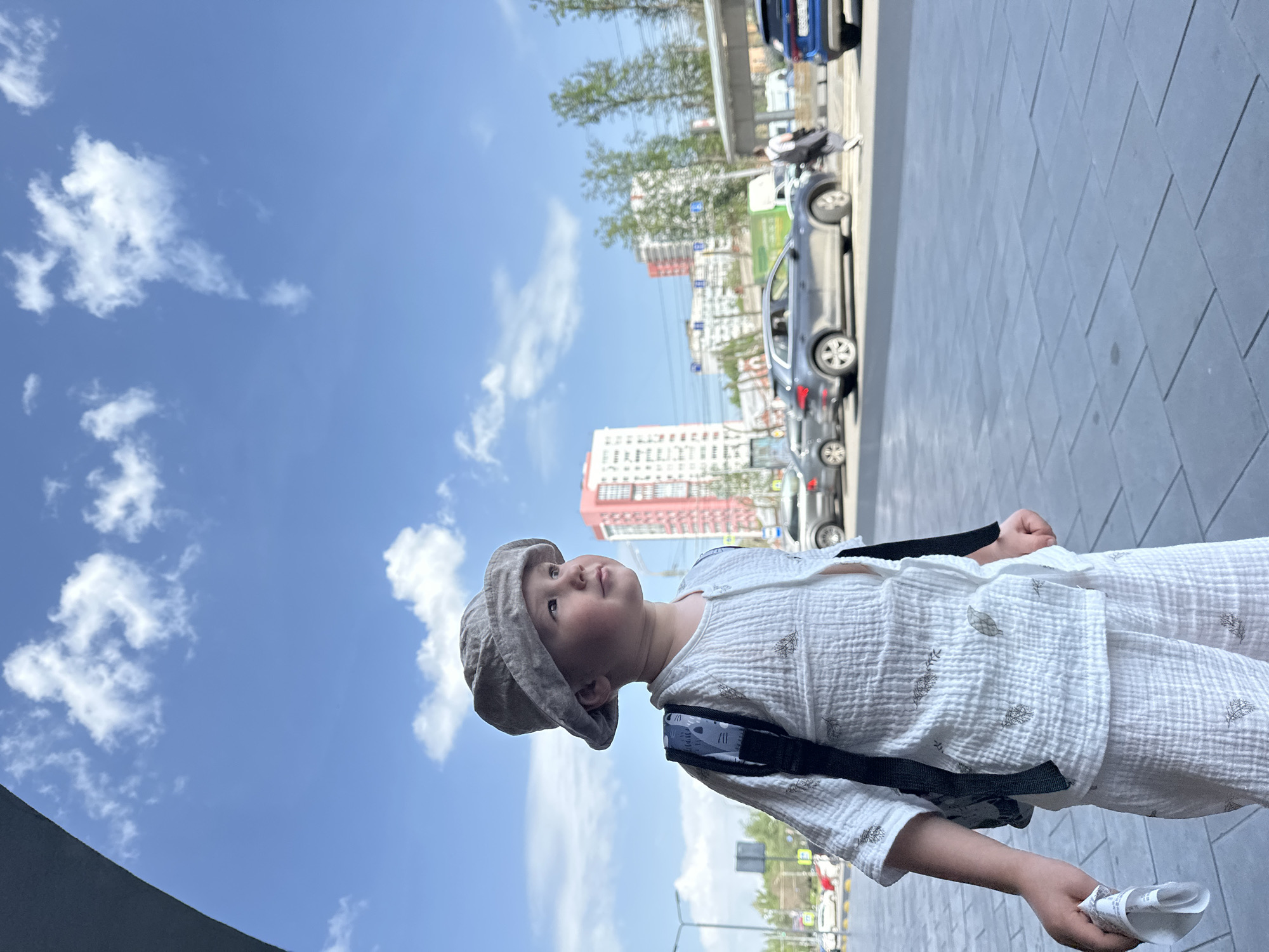 A low-angle shot depicts a young child, facing upwards and to the left, in an urban environment. The child wears a patterned white outfit and a grey hat, and has a small backpack. The backdrop features a clear, bright blue sky with scattered white clouds. In the background, there are several cars parked along the side of the road, as well as buildings, including a high-rise with a red and white design. A street with visible street signs is also in the background. Based on the architecture and urban landscape, this photo was likely taken in the city of Nizhny Novgorod, Russia.