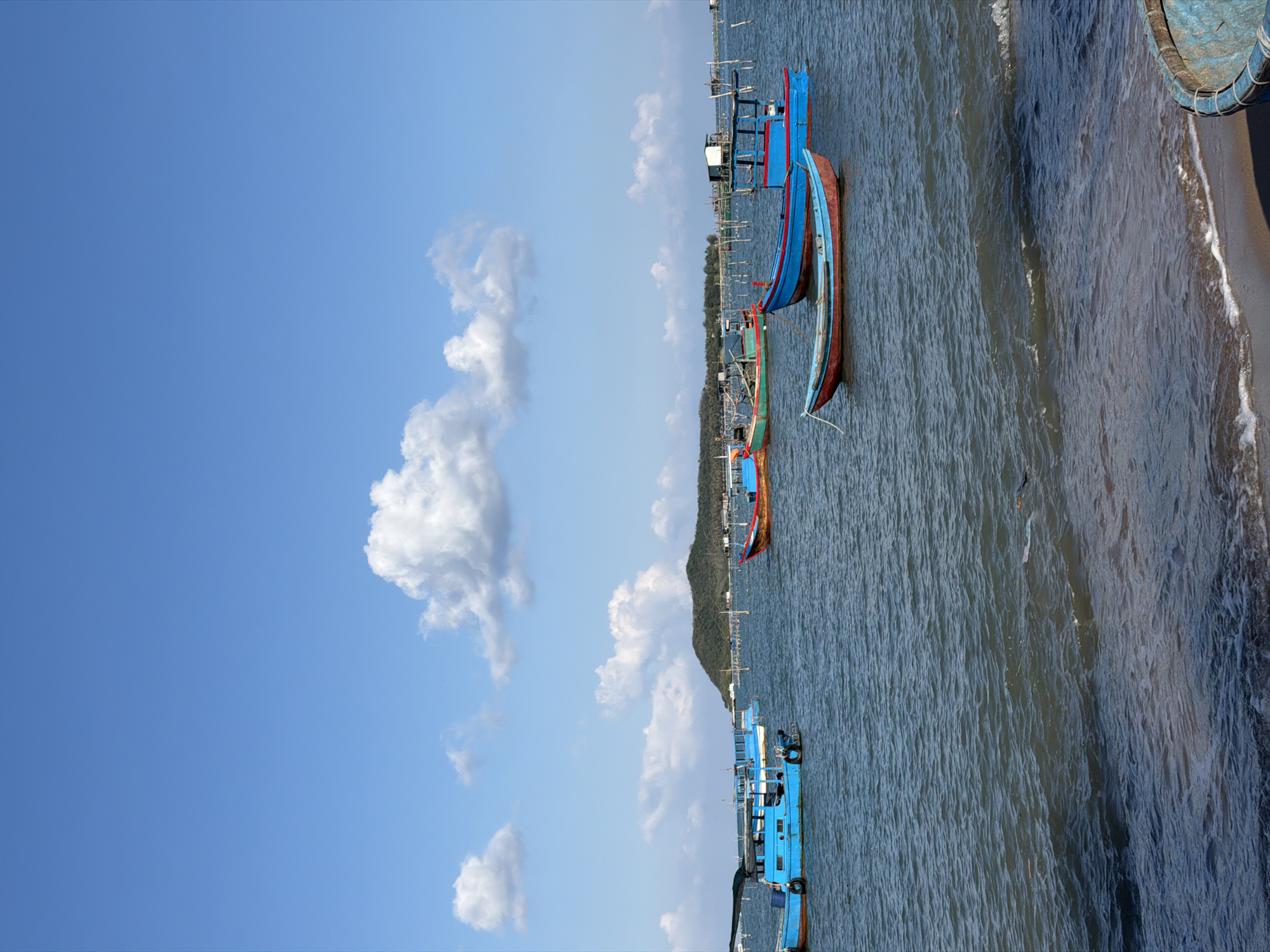 The image depicts a coastal scene in Bac Nha Trang Ward, Vietnam, featuring a row of fishing boats docked near the shoreline. The boats are painted in vibrant shades of blue and red, contrasting with the greenish-blue sea. A small strip of beach borders the water, adjacent to a sea wall. In the background, a green hillside can be seen. The sky is blue with scattered white clouds.