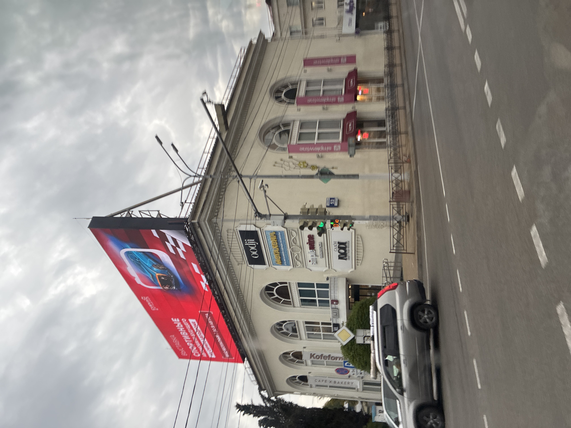 The image depicts a street scene in Sochi, Russia, under an overcast sky. A multi-story building with arched windows dominates the frame. A large, bright red billboard with an image of a blue sports car and Russian text advertising a "sports exhibition" is mounted on top of the building. Multiple businesses with signs such as "oodji," "METKHAXA," "simplewine," and "KOTOfON" are visible on the building's facade. A silver SUV is driving on the road in front of the building. Traffic lights and street lamps are also present. The scene suggests a commercial area within the city.