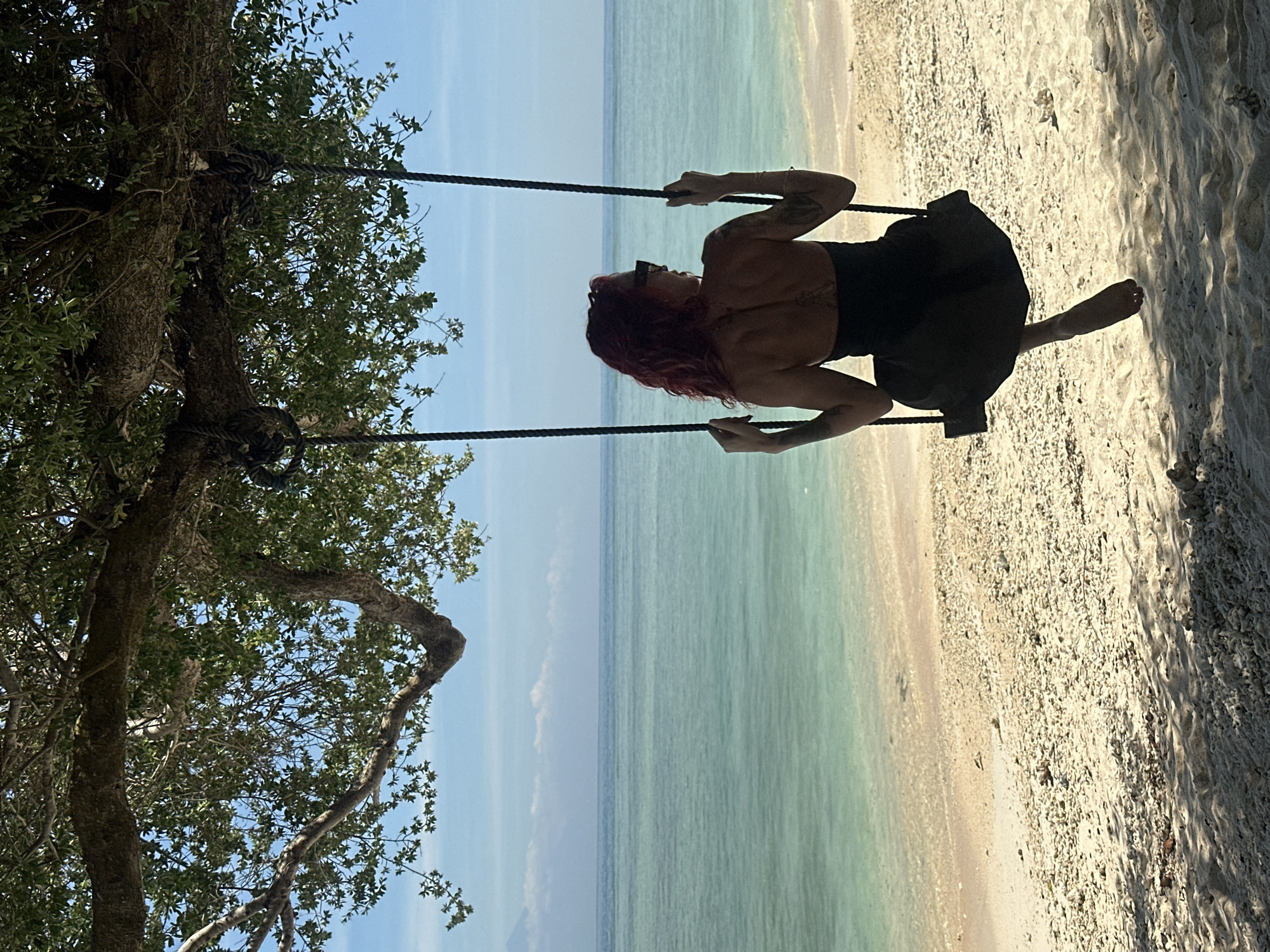 The image depicts a woman with red hair, wearing sunglasses and a dark-colored dress or top and skirt, sitting on a rope swing suspended from a tree branch on a beach. The beach borders clear, light-blue water, which merges with the sky in the background. The woman appears to be swinging. The location is Unknown, Indonesia.