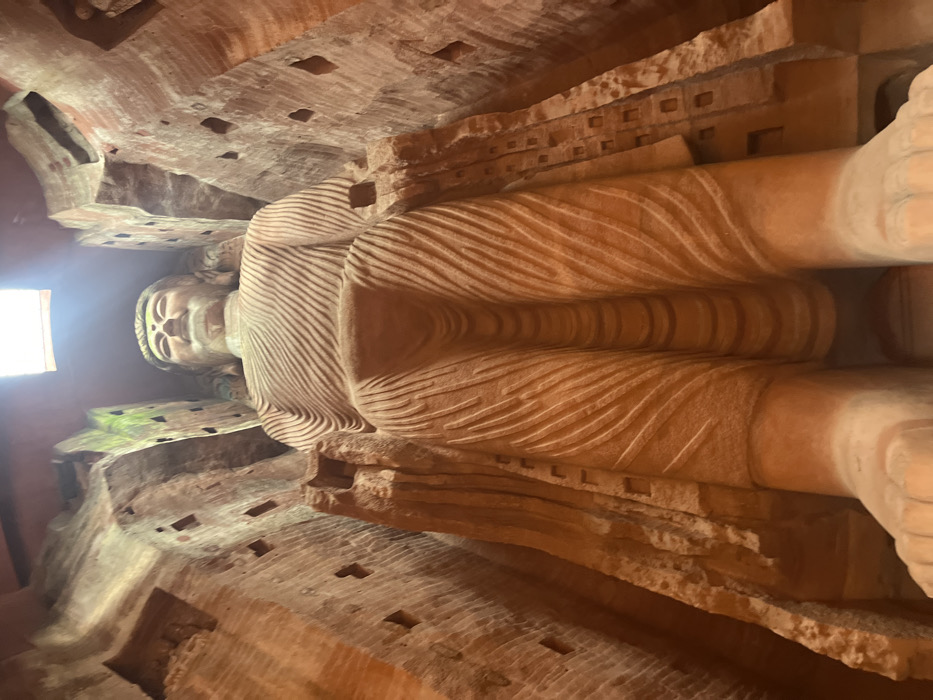 A colossal, seated Buddha statue, carved from reddish-brown sandstone, dominates the scene within a towering rock-cut niche in Shizhong District, Leshan City, China. The statue faces forward, depicted from a low-angle perspective, emphasizing its monumental scale. The Buddha features a serene expression with a downcast gaze, elongated earlobes, and a visible ushnisha. Its robes are intricately carved with numerous undulating, parallel folds, covering the body from shoulders to ankles. Portions of the feet are visible at the very bottom of the frame. The surrounding rock walls, part of the original cliff face, exhibit rough-hewn textures and contain multiple small, rectangular carved recesses. Greenish biological growth, likely moss or lichen, adheres to sections of the upper rock walls. A bright, square light source at the top center of the frame illuminates the upper half of the statue and the cave interior, casting a strong downward beam. No individuals are visible.
