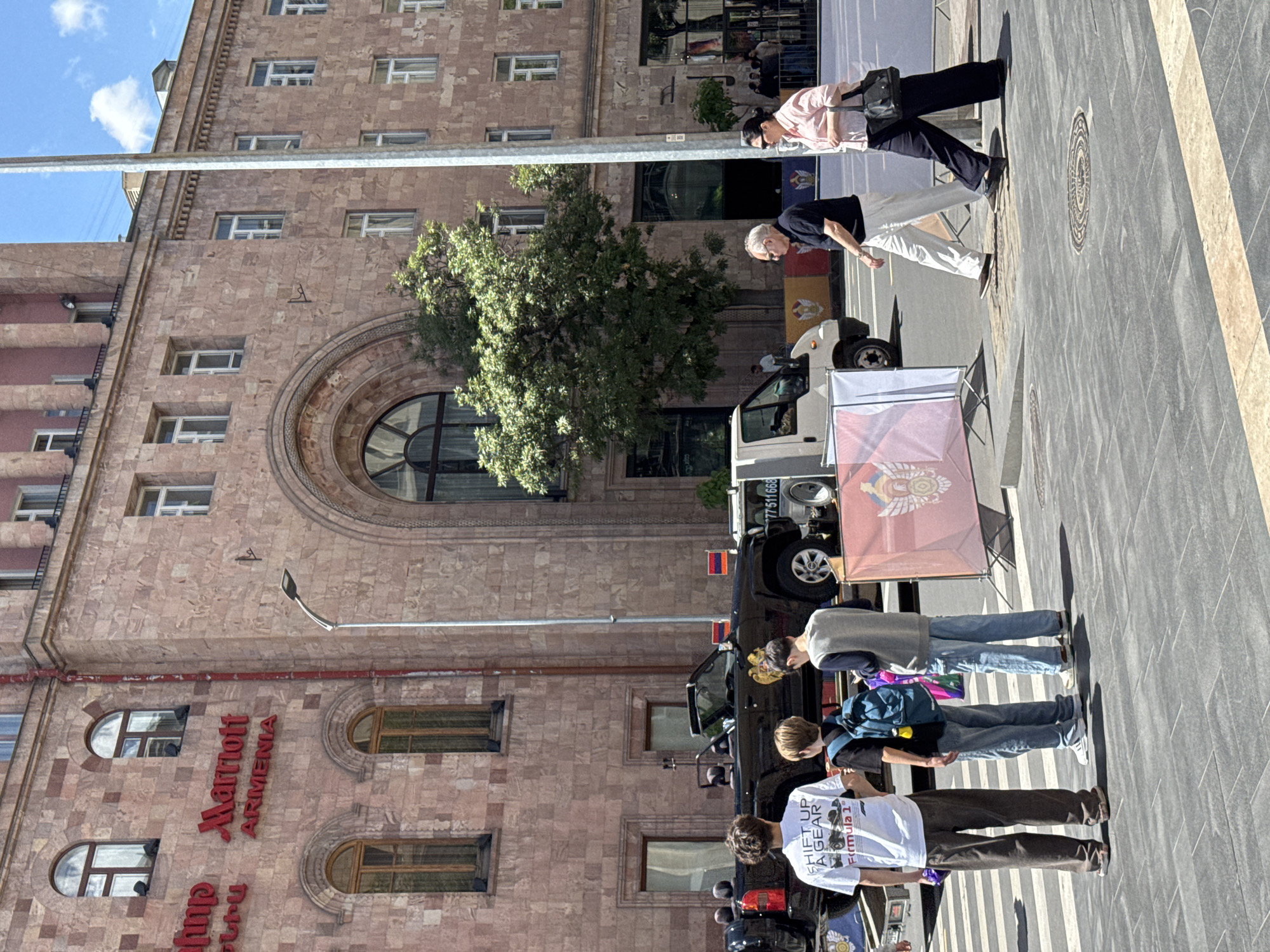 The image shows the Marriott Armenia hotel in Yerevan, Armenia. A group of people stand on the sidewalk in front of the hotel. A white van is parked nearby with a sign featuring an emblem attached. Two older adults are also visible, walking along the sidewalk towards the Marriott.