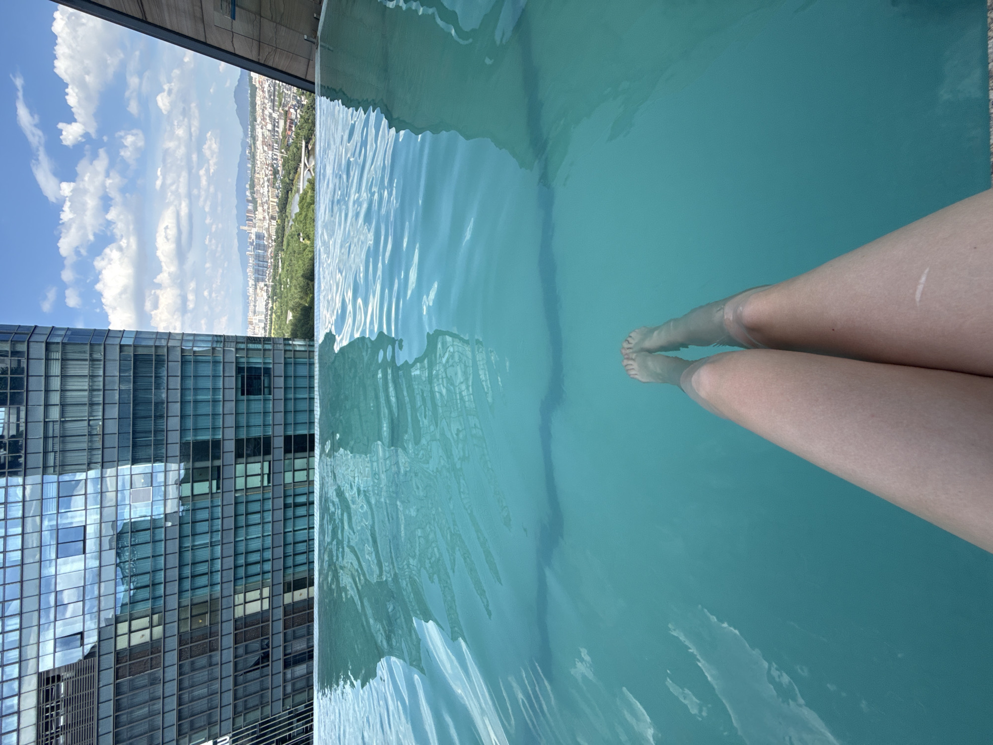 A person's legs are submerged in a bright turquoise infinity pool. The pool is positioned on a high-rise building in Yiwu, China, with a cityscape visible in the background. The cityscape features a mix of modern buildings, green spaces, and distant mountains under a blue sky with scattered clouds. The reflective facade of a tall skyscraper is prominently visible on the left side of the image.