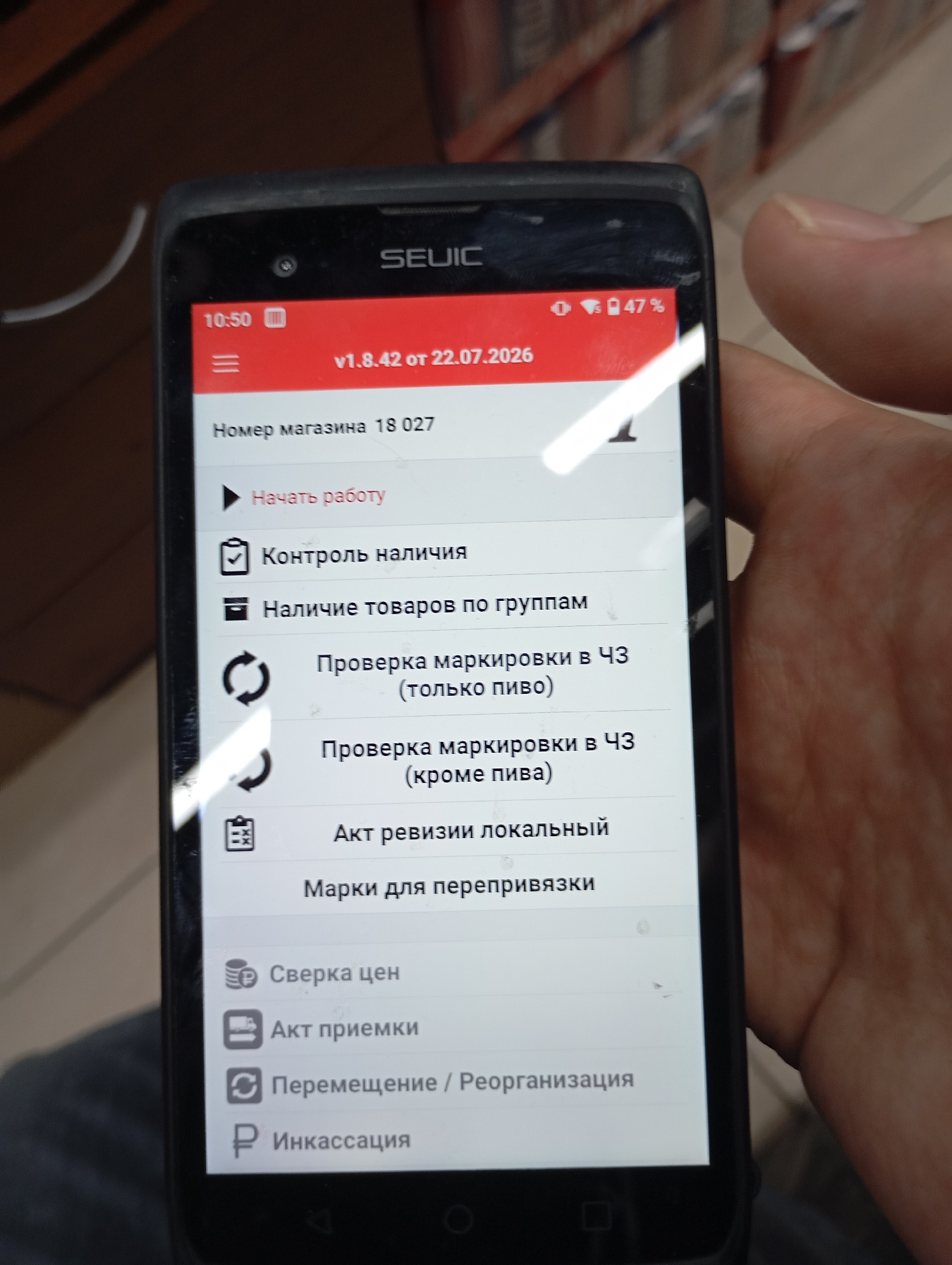 The image shows a close-up of a hand holding a SEUIC brand mobile device. The device's screen displays a Russian-language menu for a store management application. The top of the screen shows the time as 10:50, a battery indicator showing 47% charge, and the version number of the application: v1.8.42 from 22.07.2026. The main menu options visible are "Номер магазина 18 027" (Store number 18 027), "Начать работу" (Start work), "Контроль наличия" (Inventory control), "Наличие товаров по группам" (Availability of goods by groups), "Проверка маркировки в ЧЗ (только пиво)" (Marking check in CZ (beer only)), "Проверка маркировки в ЧЗ (кроме пива)" (Marking check in CZ (excluding beer)), "Акт ревизии локальный" (Local audit report), "Марки для перепривязки" (Marks for re-binding), "Сверка цен" (Price reconciliation), "Акт приемки" (Acceptance certificate), "Перемещение / Реорганизация" (Movement / Reorganization), and "Инкассация" (Cash collection). The background is blurred and appears to be an indoor environment, possibly a retail store. There are no visible landmarks or clues that would indicate the specific location within Moscow, Russia. The image is a still photograph taken from a first-person perspective.
