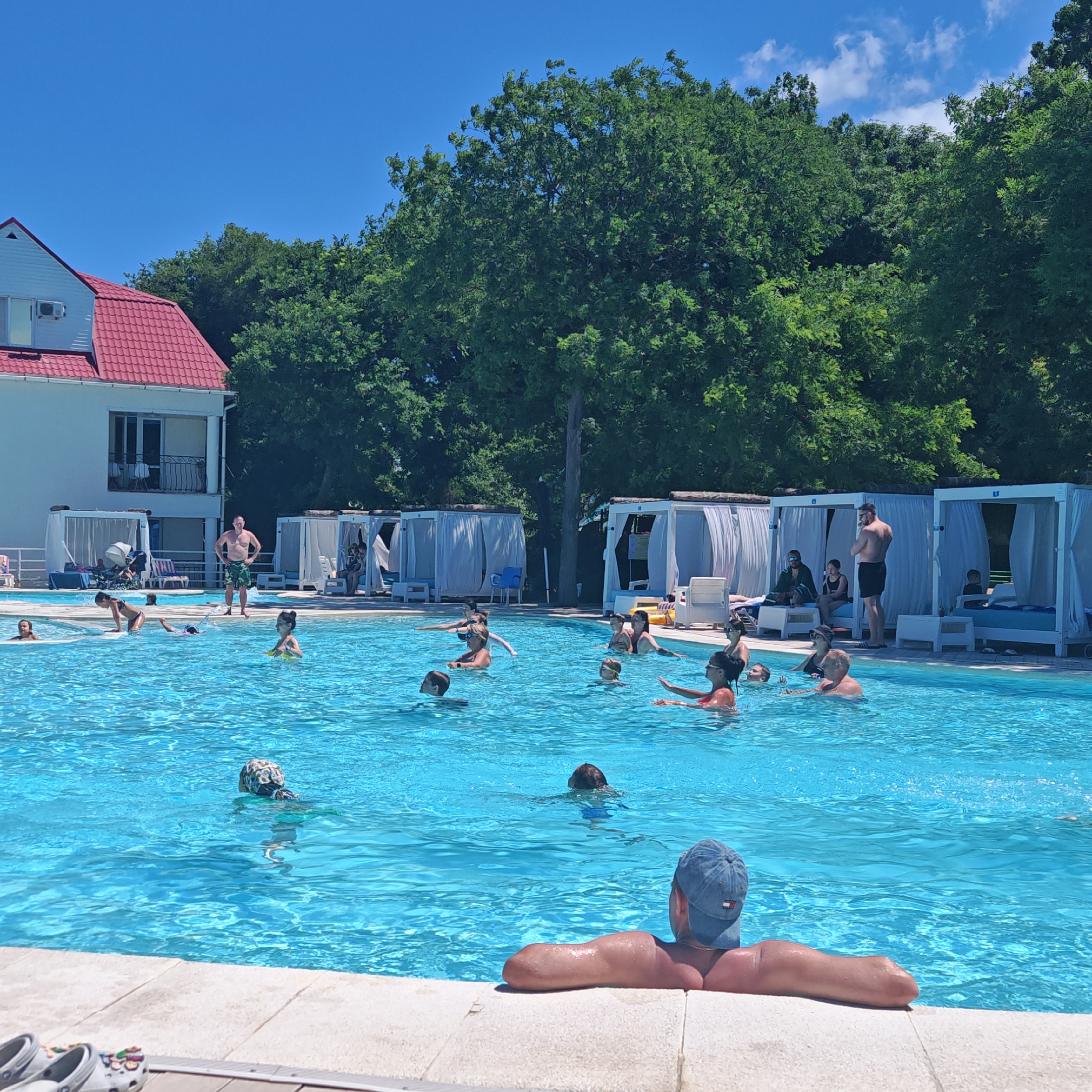 The image depicts an outdoor swimming pool area filled with people enjoying a sunny day. The pool is a large, bright blue expanse of water, with several individuals swimming and interacting. In the foreground, a person wearing a blue baseball cap is seen from the back, resting their arms on the pool's edge. Further into the pool, various individuals are engaged in activities such as swimming, playing, and what appears to be a water aerobics class, with several participants performing synchronized arm movements. Some people are seen with swimming caps and goggles.
Behind the pool, a row of white cabanas with draped curtains offers shaded seating areas. Some people are seated within or near these cabanas. A white building with a distinctive red tiled roof is visible on the left, with balconies and windows, suggesting it is part of a resort or hotel. Lush green trees form a dense backdrop behind the cabanas and building. The sky is clear and blue, indicating fair weather. The overall scene suggests a recreational setting, likely a hotel or resort pool in a location that experiences warm weather, consistent with a place like Gorshkove (городское поселение Щелкино), Ukraine. The image captures a lively atmosphere of people enjoying leisure time by the water.