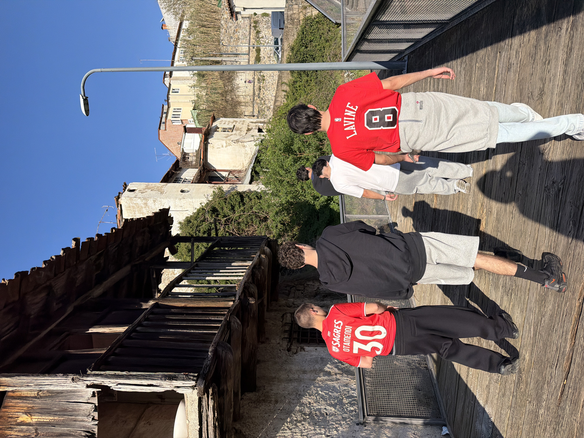 The image depicts six individuals walking away from the camera on a wooden walkway in a historic section of Bragança, Portugal, under a clear blue sky.
On the immediate left, an old two-story stone building with a weathered wooden balcony and a tiled roof is prominent. The timber of the balcony appears dark and aged. Behind this, lush green vegetation and additional older buildings with light-colored facades and tiled roofs extend up a hillside.
The walkway itself is constructed from horizontal wooden planks and features a grey metal mesh railing on its right side. Beyond this railing, a tall, curved grey streetlamp stands. Further into the background on the right, a terraced hillside reveals a densely built urban environment with various residential buildings, including some with light-colored walls and red-tiled roofs. A stone retaining wall is visible, and a small, distant human figure can be seen on an upper path. Partially visible vehicles are present on a lower road.
The six individuals are all male, casually dressed, and walking in unison. The person on the far left wears a red short-sleeved jersey with "SAGRES OTAMENDI 30" printed on the back and black trousers. Next to him, a person wears a black hooded sweatshirt, grey shorts, and black athletic shoes. The person on the far right wears a red short-sleeved shirt with "LAVINE 8" on the back, light blue jeans, and a grey, skirt-like garment worn over the jeans. Three additional figures are partially visible walking behind them. Strong sunlight, likely from the late afternoon or early morning, casts long, defined shadows of the individuals and the railing across the wooden walkway.
