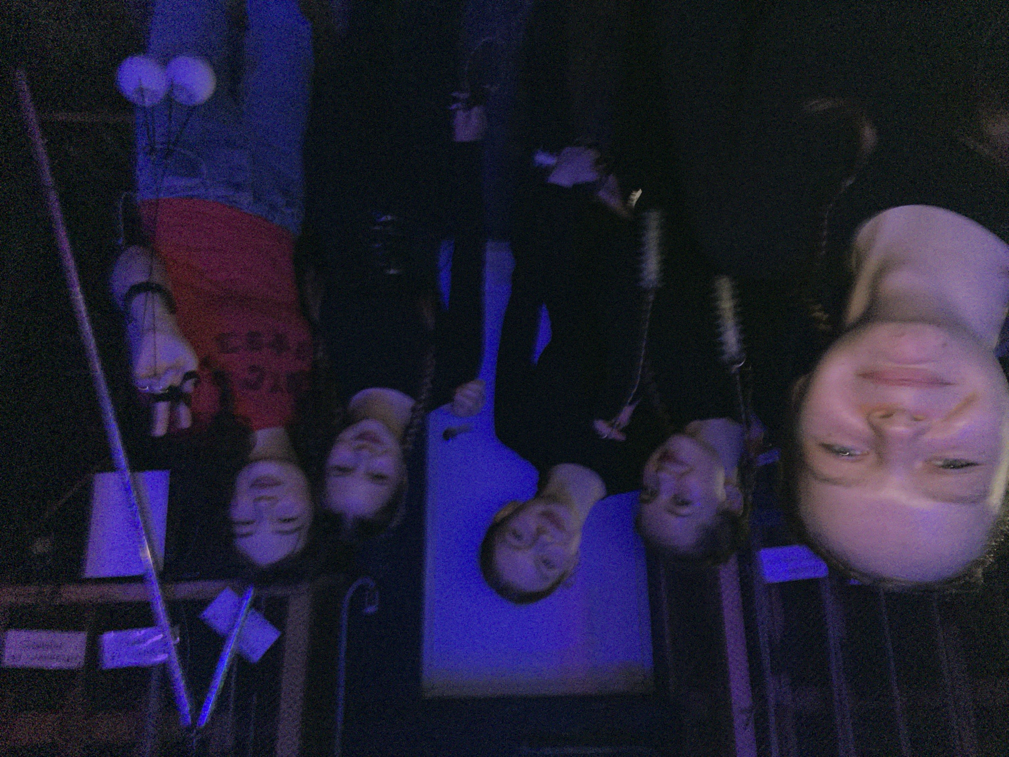 In a dimly lit room with purple lighting, five young females are posing for a selfie. The person in the foreground on the left is a blonde female with her mouth slightly open, looking towards the camera. To her right are three other females, all smiling and looking at the camera. The female furthest to the right is wearing a red shirt and blue jeans, and is holding what appears to be a pair of poi balls. The background is dark and appears to be a room with some scaffolding or stage equipment, possibly in a community center or performance space in Chaikovsky, Russia.