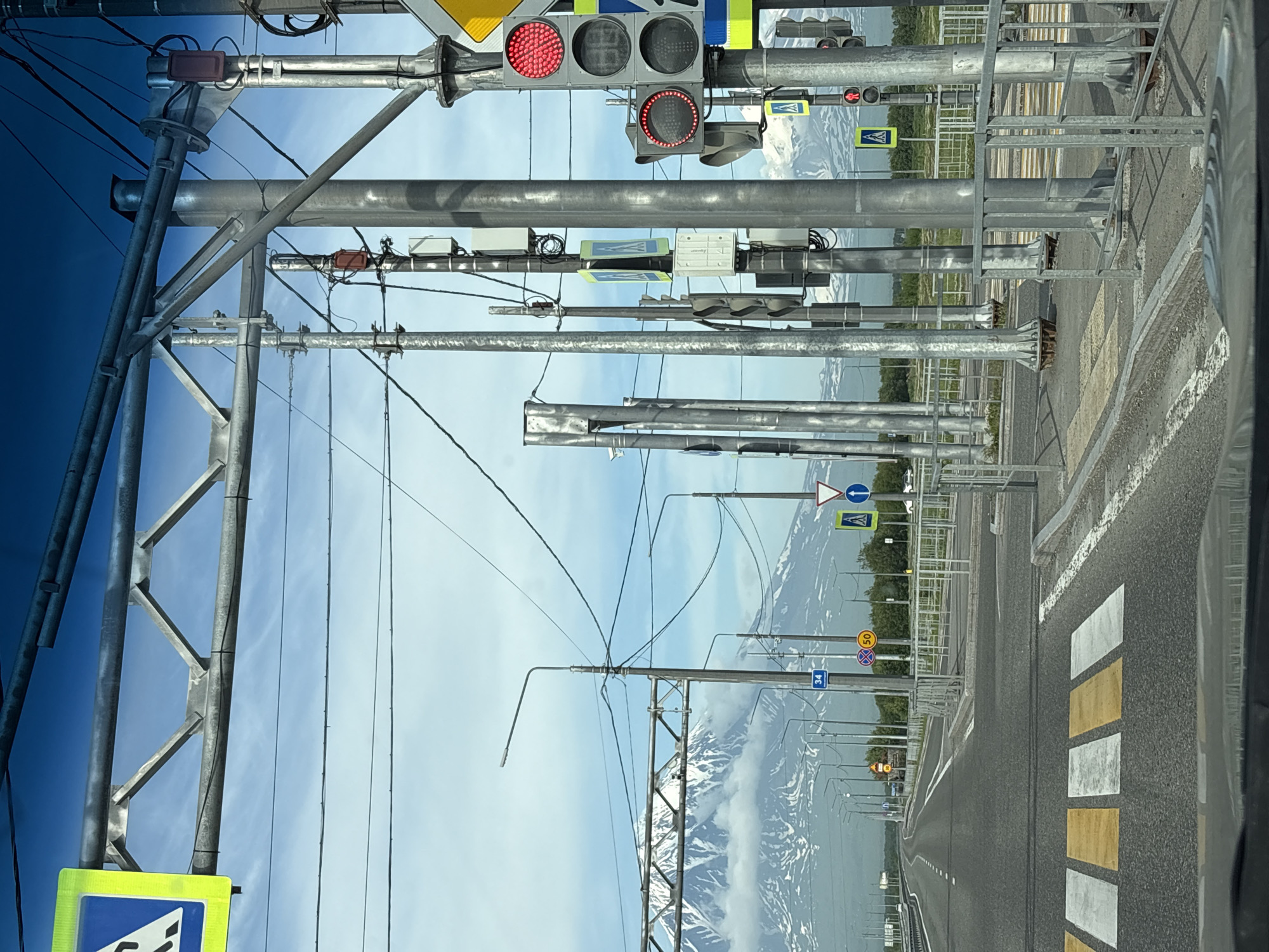 The image depicts a view from a vehicle on a road in Yelizovo, Russia, with a prominent snow-capped mountain range in the background, likely Mount Avachinsky or Mount Koryaksky. The foreground shows a road with a pedestrian crossing marked by yellow and white stripes. Several traffic light poles are visible, equipped with traffic signals, road signs, and communication equipment. Some traffic lights are illuminated red. The road appears to be an asphalt highway or main road, with multiple lanes. Fencing lines the sides of the road. Overhead power lines and metal support structures are also present. The sky is partly cloudy.