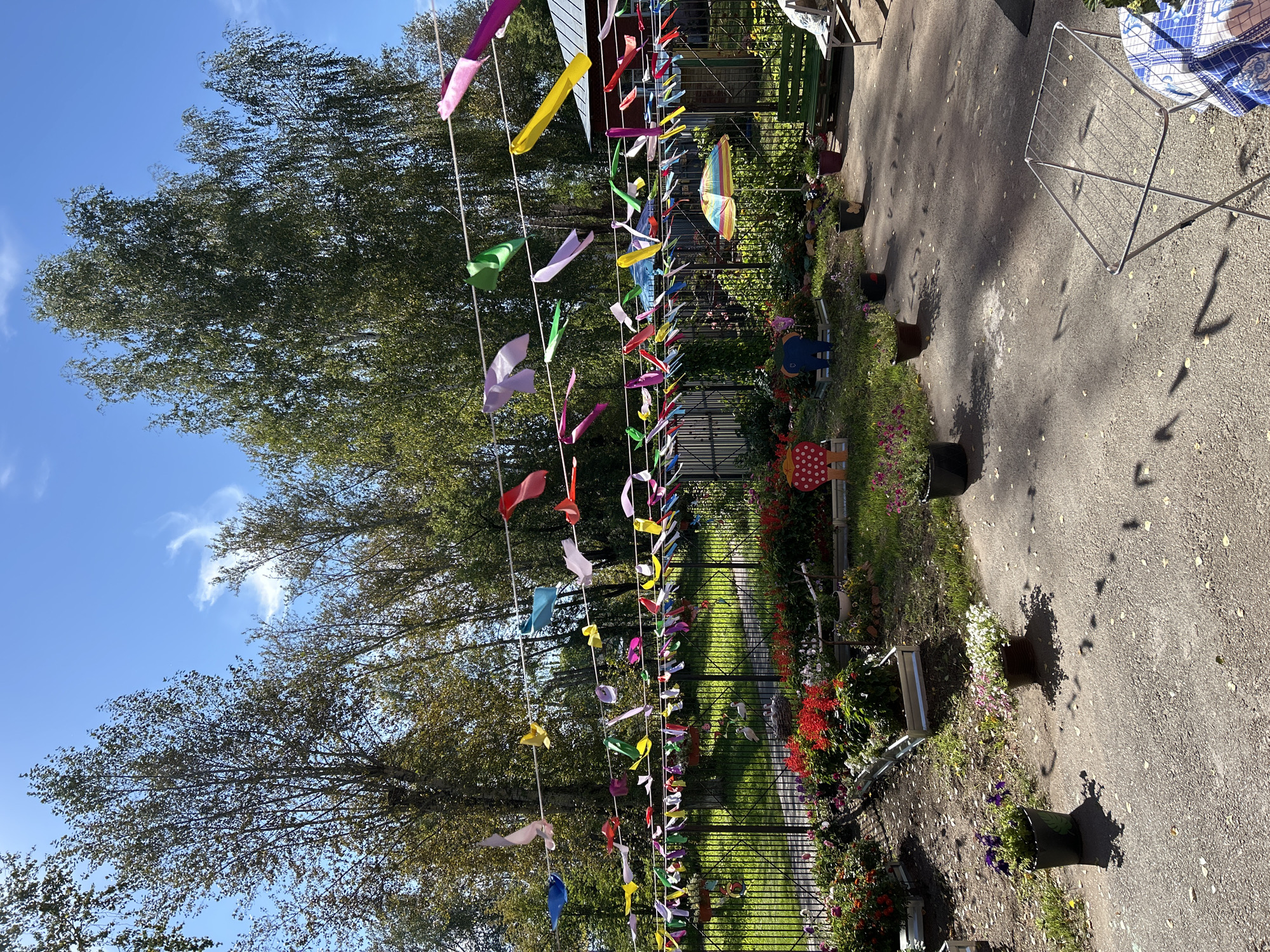 The image depicts a sunny outdoor scene, likely a backyard or playground, decorated with colorful streamers strung between trees and a fence. The streamers are a variety of colors and shapes, fluttering in the breeze. Below the streamers, a black metal fence encloses a garden area filled with various flowers, predominantly red and pink. There are also several decorative items visible, including a small umbrella, a blue elephant figurine, and a wooden cutout of a ladybug. In the foreground, a paved area has shadows cast by the trees and streamers. A collapsible drying rack stands on the pavement. In the background, trees and a building are visible. The location is stated to be the city of Tomsk, Russia. There are no people visible in the image.