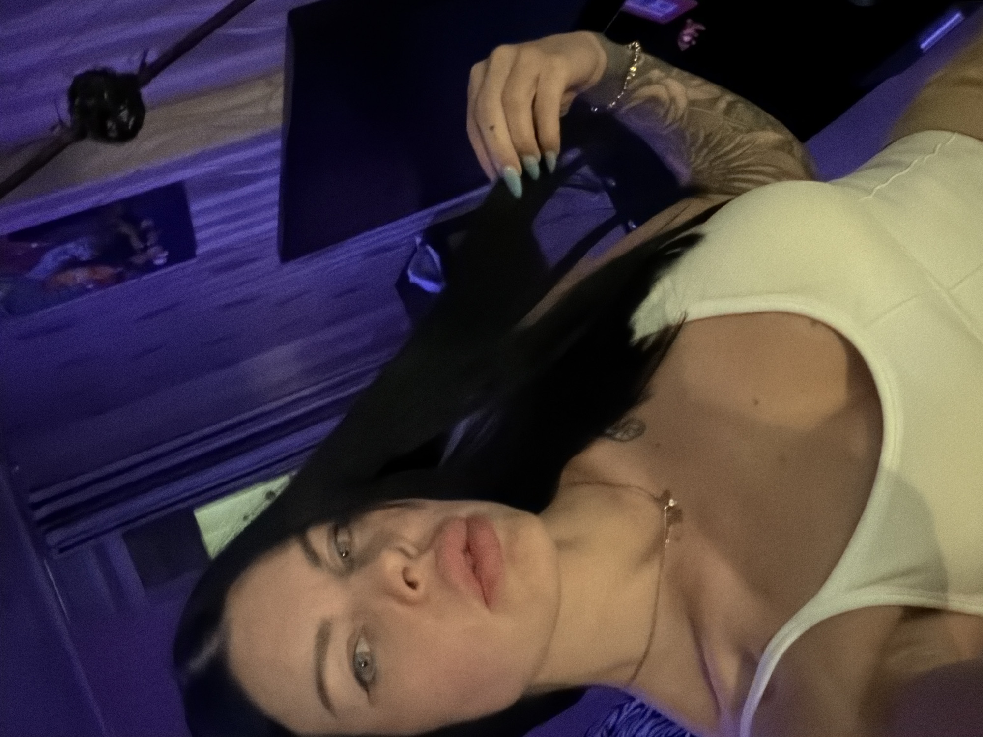 The image shows a selfie of a woman with long, dark hair and light-colored eyes, wearing a white top. She has visible tattoos on her arm and a necklace around her neck. The background appears to be an indoor setting with purple lighting and curtains. There are no discernible landmarks or indicators to suggest the location is Ufa, Russia.