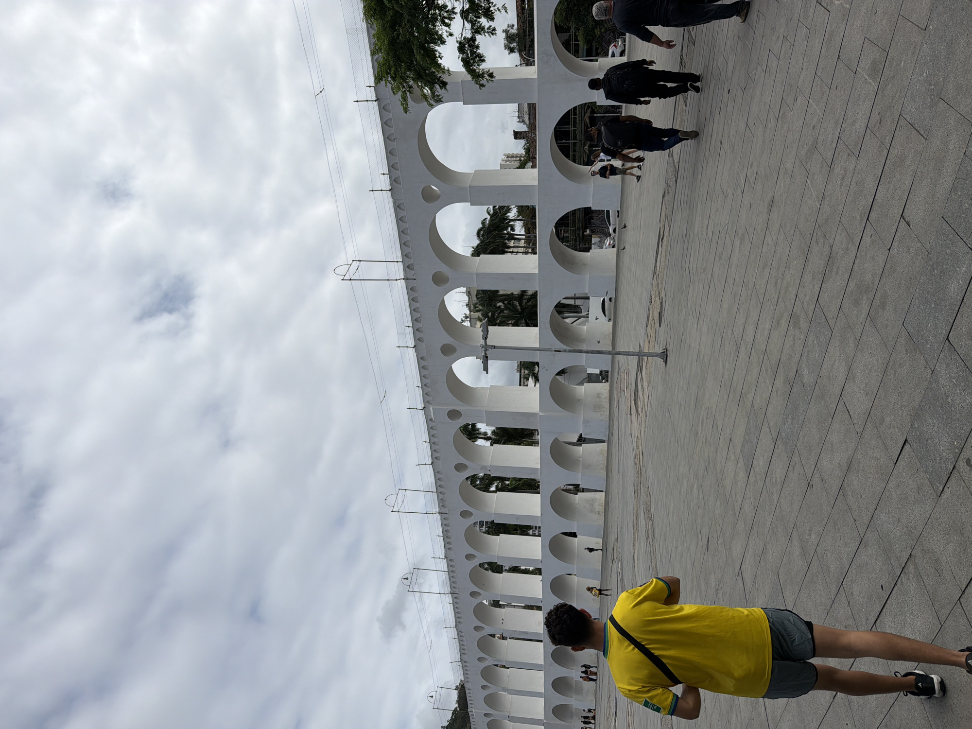 The image shows the Arcos da Lapa in Rio de Janeiro, Brazil, on an overcast day. In the foreground, a young man wearing a yellow t-shirt and grey shorts is walking away from the camera. Behind him, several other people are walking across a paved plaza in front of the aqueduct. The aqueduct itself is a long, white structure with multiple arches, and in the background, there are buildings and trees. The sky is filled with clouds.
