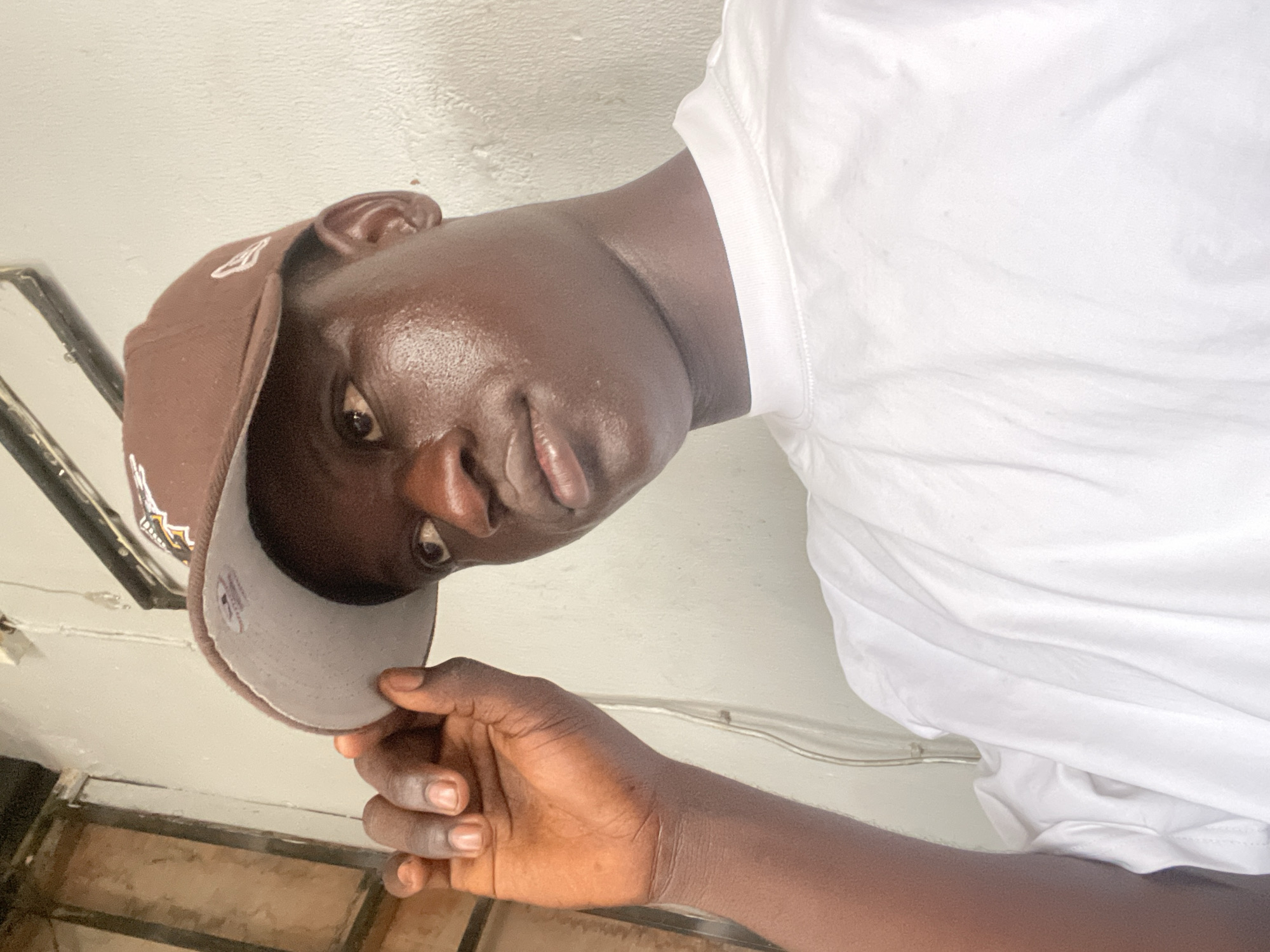 A low-angle close-up shot of a dark-skinned man wearing a brown baseball cap and a white t-shirt. The man is holding the brim of his cap with his left hand. The location appears to be indoors, possibly in a room with white walls. Given the prompt includes the city of Ado, Nigeria, it is implied the subject is likely in that location. However, there are no details in the image to independently verify the geographic location.