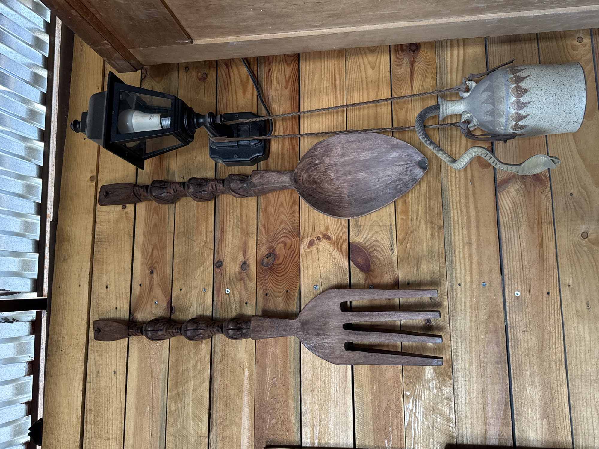 This image presents a rustic wall decoration in Yerevan, Armenia, featuring various items mounted on vertical wooden planks. The wooden wall exhibits natural grain, knots, and visible screws. On the upper left, a black, glass-paneled lantern, typical of outdoor fixtures, is mounted, housing a white LED light bulb with "PARMIDA dimmable" text partially visible on its base. A black electrical wire extends from the lantern. Spanning across the wall are two twisted ropes or cords. Below the lantern, a large, dark brown wooden spoon with an ornate, carved handle is displayed horizontally. Below that, a similarly large wooden fork with a matching carved handle is mounted. To the right, a light-colored ceramic jug or vase is suspended by leather-like straps. The jug features a subtle patterned decoration of dark, inverted triangular shapes. A light green, realistic-looking serpentine figure, likely a decorative snake made of rubber or plastic, is draped around the jug, with its head poised upwards. One of the twisted ropes connects the lantern's mounting area to the jug, while the other runs below, appearing to link the spoon's handle to the jug. On the far left, a corrugated metal surface is partially visible. The scene is a static display, suggesting a decorative arrangement in a home or establishment.