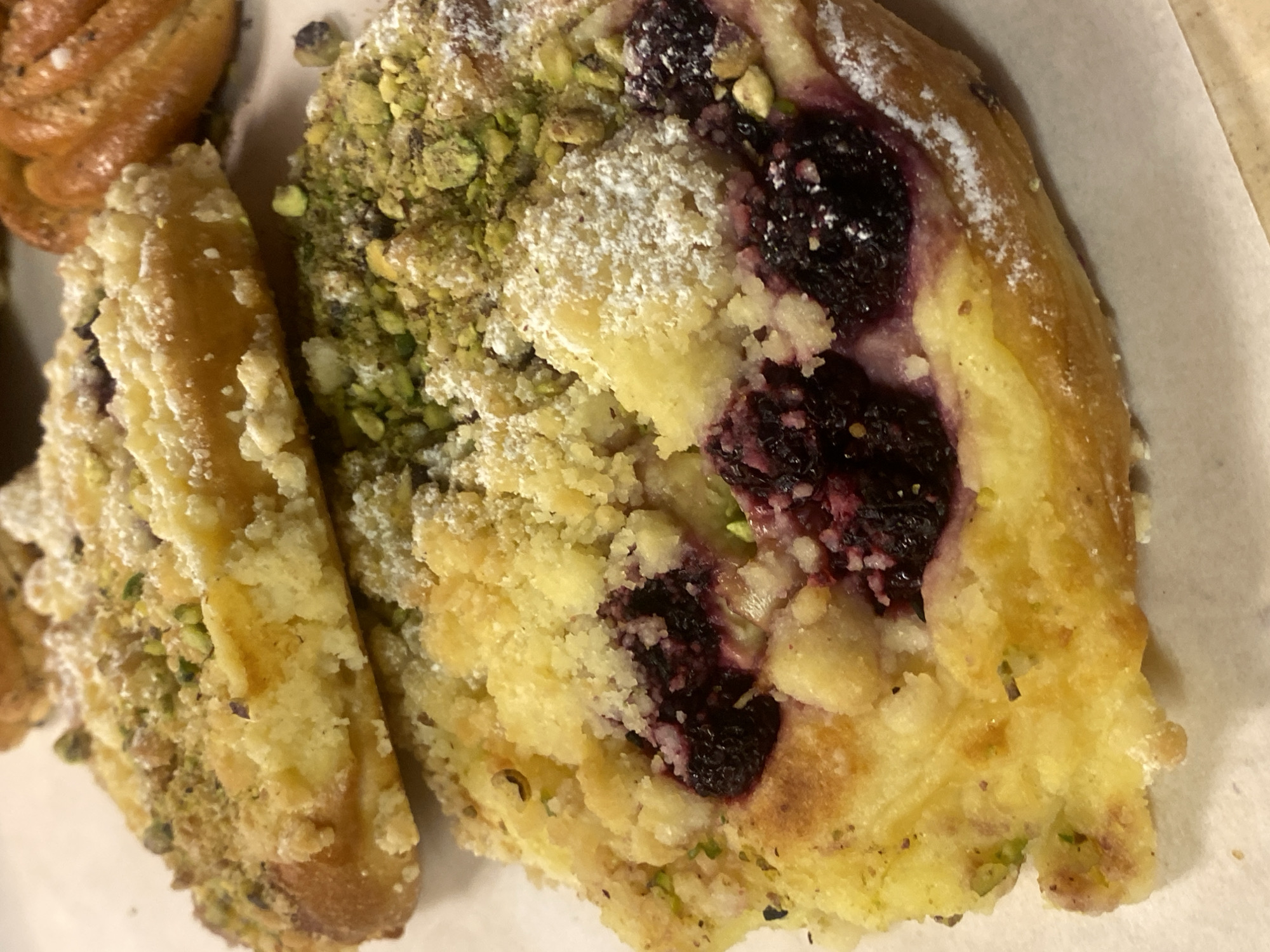 The image displays a close-up of a pastry, likely a type of fruit danish or tart, with a prominent filling of dark berries, possibly blueberries or blackberries. The pastry has a golden-brown, flaky crust. A crumb topping, a mixture of crumbled dough and finely chopped pistachios, covers a significant portion of the pastry. The entire pastry is lightly dusted with powdered sugar. The pastry is positioned on a white surface, possibly a baking sheet lined with parchment paper. In the background, another pastry, with a swirled shape and a similar golden-brown color, is partially visible. The lighting suggests it might be an indoor setting, with the focus solely on the food item. The colors are warm, highlighting the rich tones of the pastry and berries, with the vibrant green of the pistachios providing a contrast. There is no visible text in the image.