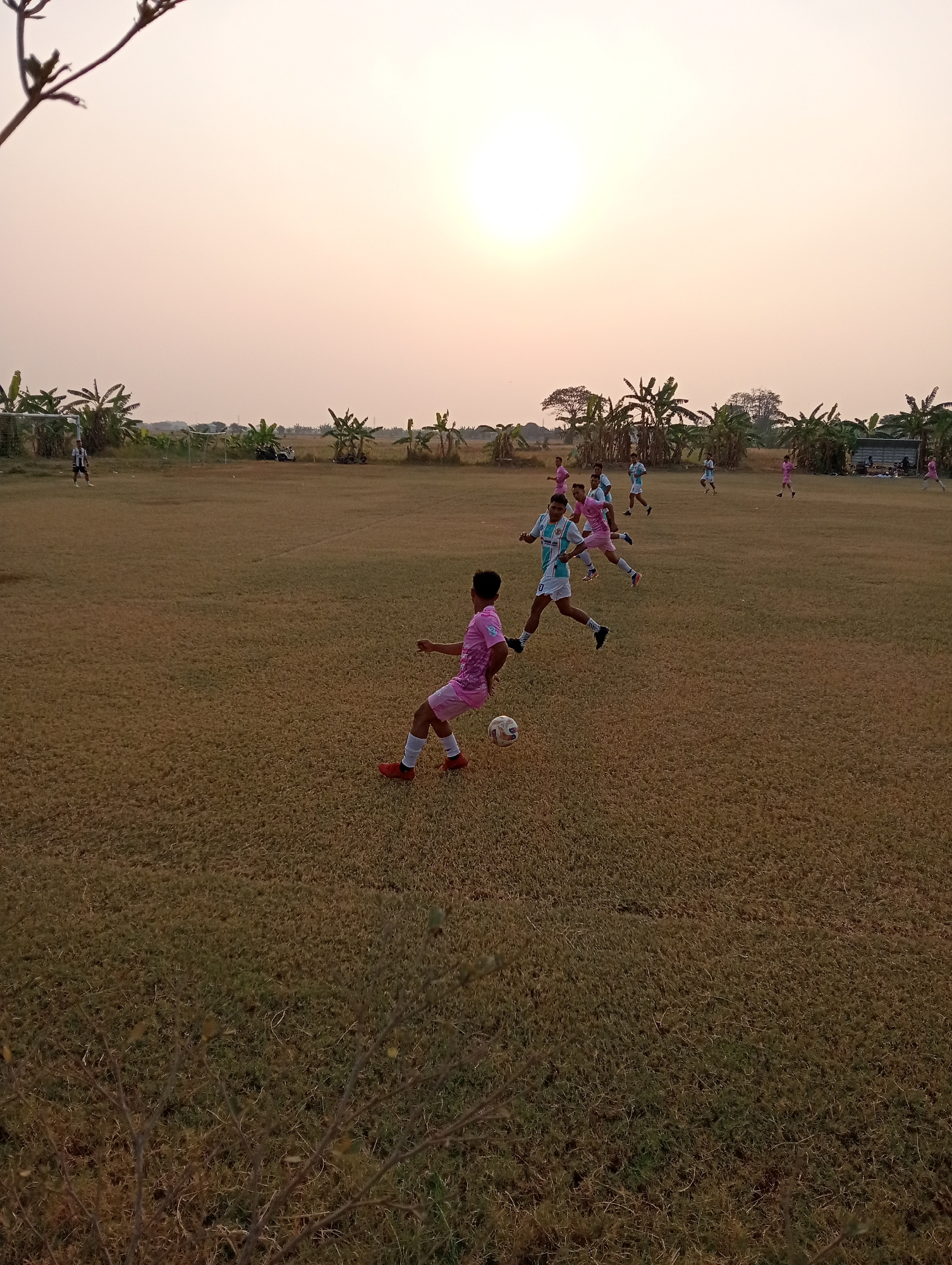 The image depicts a soccer game taking place on a dry, grass field, likely in the city of Kenep, Indonesia, during the late afternoon or early evening, indicated by the warm, diffused sunlight and the low angle of the sun in the sky. Several individuals, presumed to be players, are scattered across the field, engaged in the game. One player in the foreground, wearing a pink jersey, is in the motion of kicking a soccer ball. Other players, in pink and light blue jerseys, are also visible, some in pursuit of the ball or positioned on the field. The background is characterized by a line of trees and vegetation, possibly banana trees, and a hazy sky with the sun visible. Two soccer goals are present on the field, one on the left and another further back. The overall impression is that of a casual soccer match in a rural or semi-rural outdoor setting.