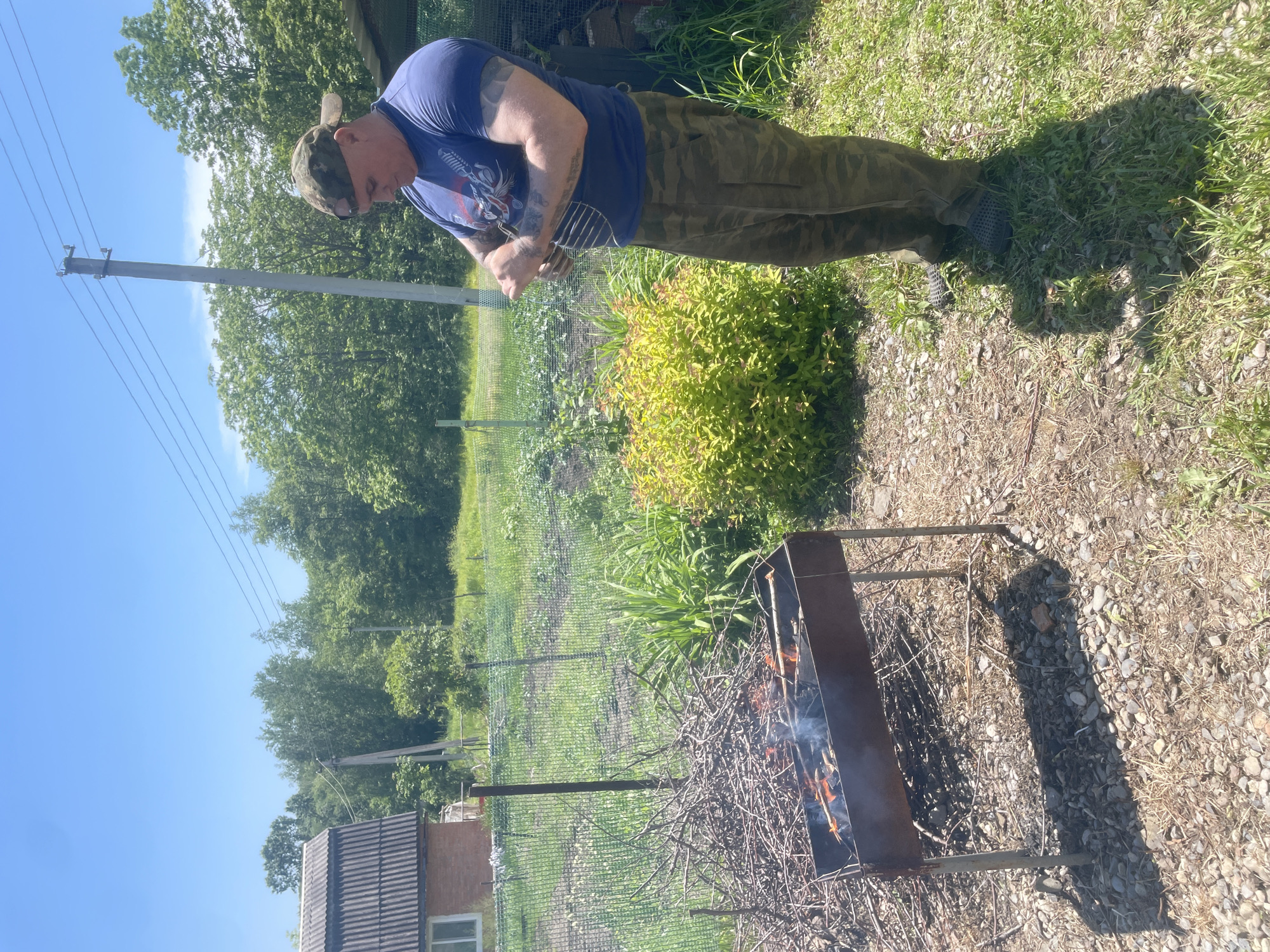 In the city of Yasnoe, Russia, a man wearing a camouflage cap, a blue t-shirt with a graphic print, and camouflage pants stands in a yard preparing to grill. He is holding a metal grill rack, and his arms are covered in tattoos. In the foreground, a rusty barbecue grill is set up with a fire burning inside, and several sticks with food items are placed over the flames. To the left of the grill, a pile of dry branches is stacked. The background shows a green lawn, a fence, a building with a corrugated roof, and lush trees under a clear blue sky. The ground is covered with pebbles and dry grass.