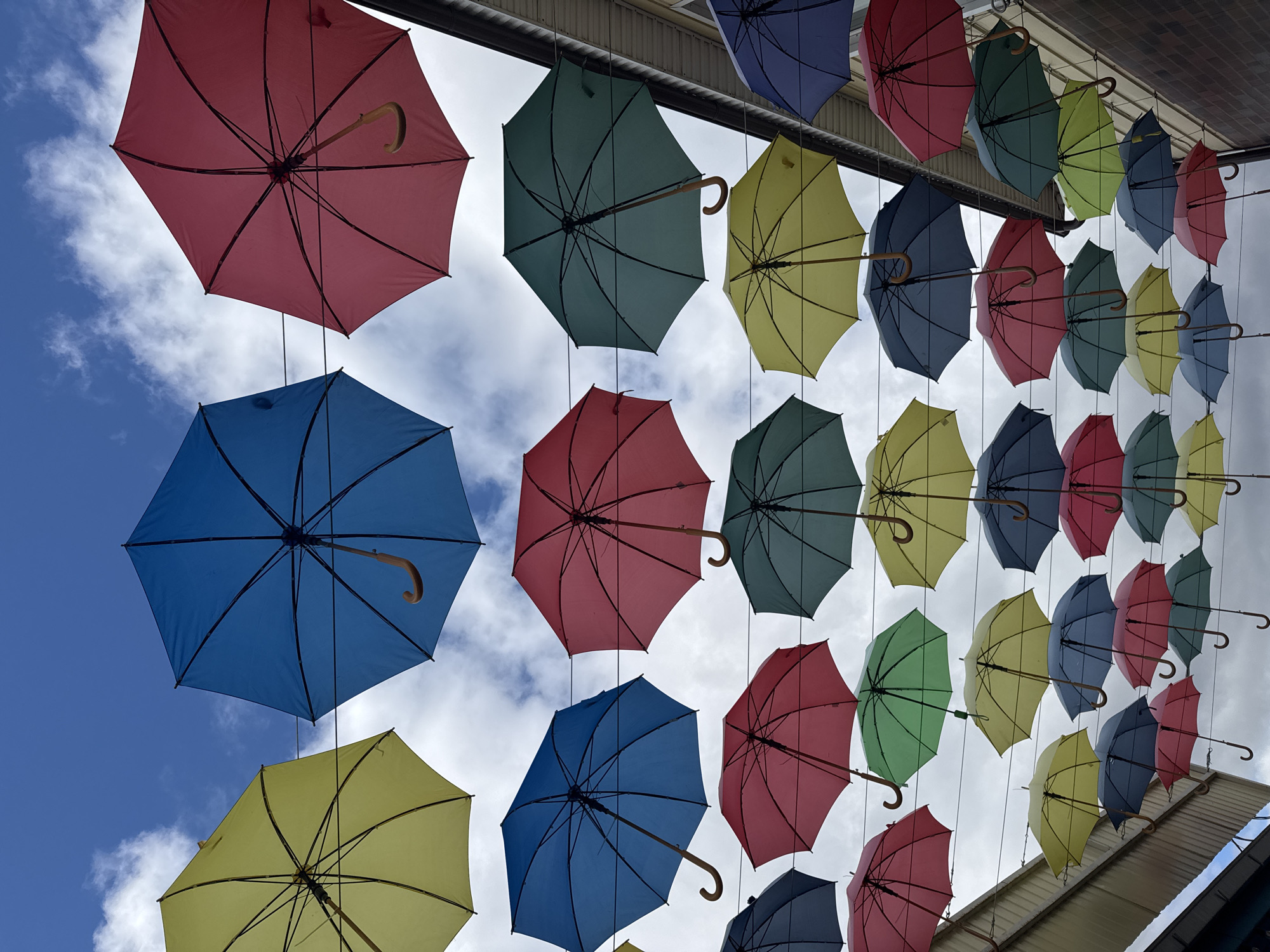A ground-level view captures rows of colorful, open umbrellas suspended above, creating an overhead canopy. The umbrellas, in shades of red, blue, green, and yellow, hang from a structure that appears to be part of a building or awning. Glimpses of the sky, including patches of blue and some white clouds, are visible between the umbrellas. The scene seems to be a street decoration. The location is likely Osa, Russia.