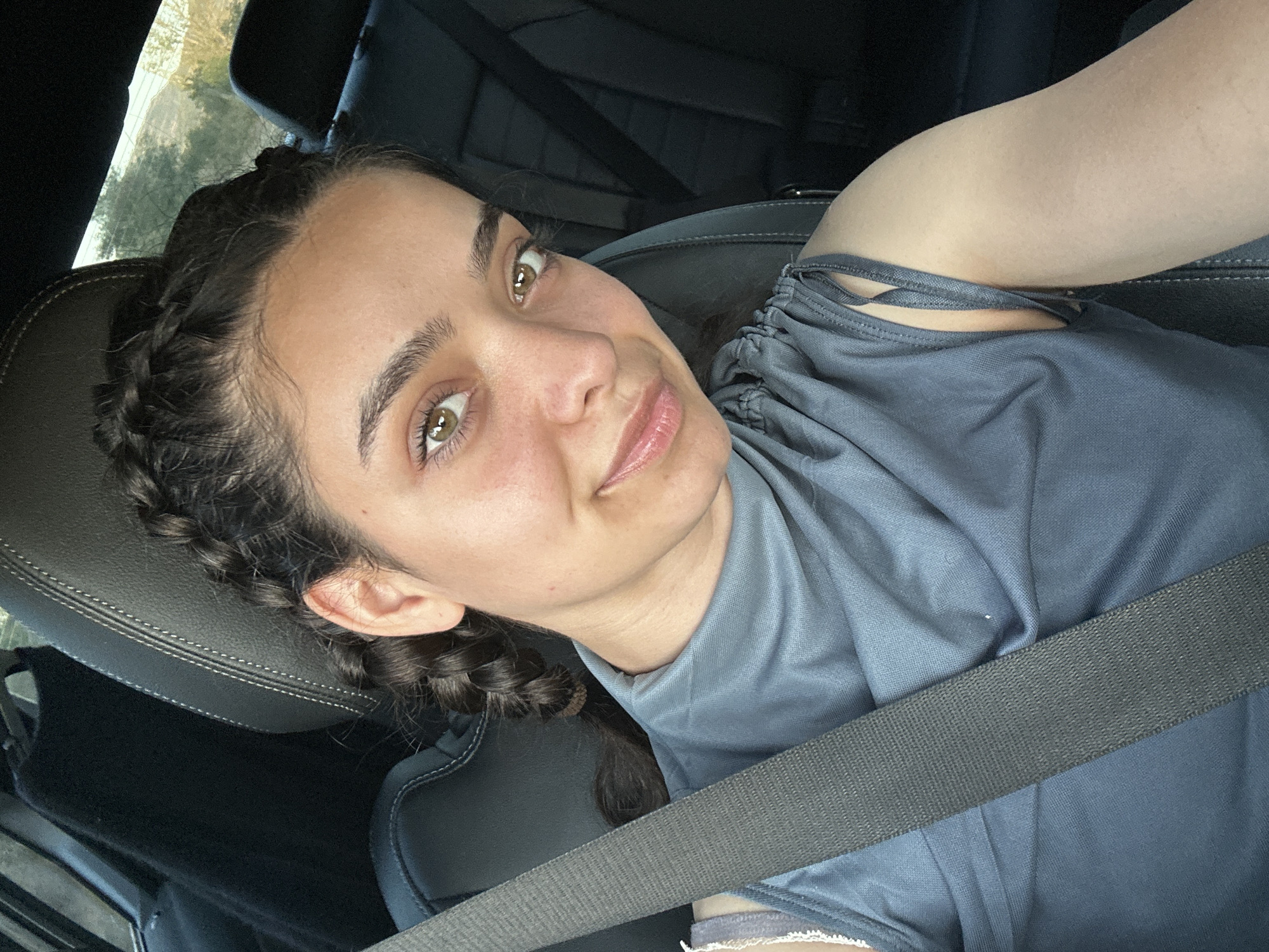 A young woman with dark hair in braids is seated in a car, wearing a gray shirt and a seatbelt. The image appears to have been taken in Drianova Glava, Bulgaria, based on context. The woman is looking towards the camera with a slight smile. The interior of the car is visible, with black leather seats and seatbelts. The background suggests an outdoor environment with trees and hills.