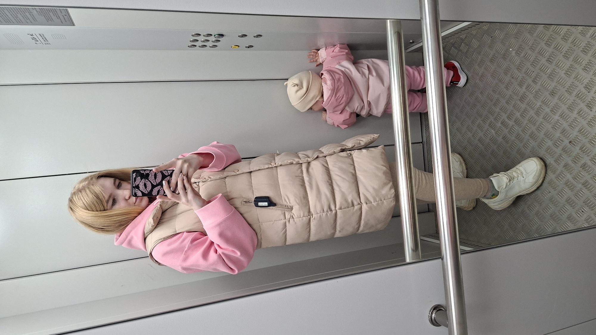 This image captures a mirror selfie taken inside an elevator, likely in Chaikovsky, Russia. The setting features a reflective wall, along with light gray solid walls, a control panel with multiple buttons (some illuminated), and a textured metal floor. Vertical stainless steel handrails are also visible, running from floor to ceiling.
The primary subject is a young woman with blonde hair, who is taking the photograph. She is dressed in a pink hooded sweatshirt, layered under a beige sleeveless puffer vest, and wears light-colored pants or leggings with chunky white sneakers. She holds a smartphone with a distinctive black case adorned with pink lips. Her expression is calm and neutral as she looks into the phone's camera.
Beside her, on the elevator floor, a small child, likely a baby or toddler, is lying down. The child is dressed in warm pink clothing, possibly a snowsuit, and a light beige beanie. One foot, wearing a red shoe, is visible. The child appears to be looking towards the back of the elevator.
The activity depicts a casual, everyday moment of a mother with her child in an elevator. The warm clothing of both individuals suggests cool weather. Above the elevator control panel, a white rectangular sign with some faint, unreadable text is present. The overall scene provides a candid snapshot of daily life within the confined space of an elevator.