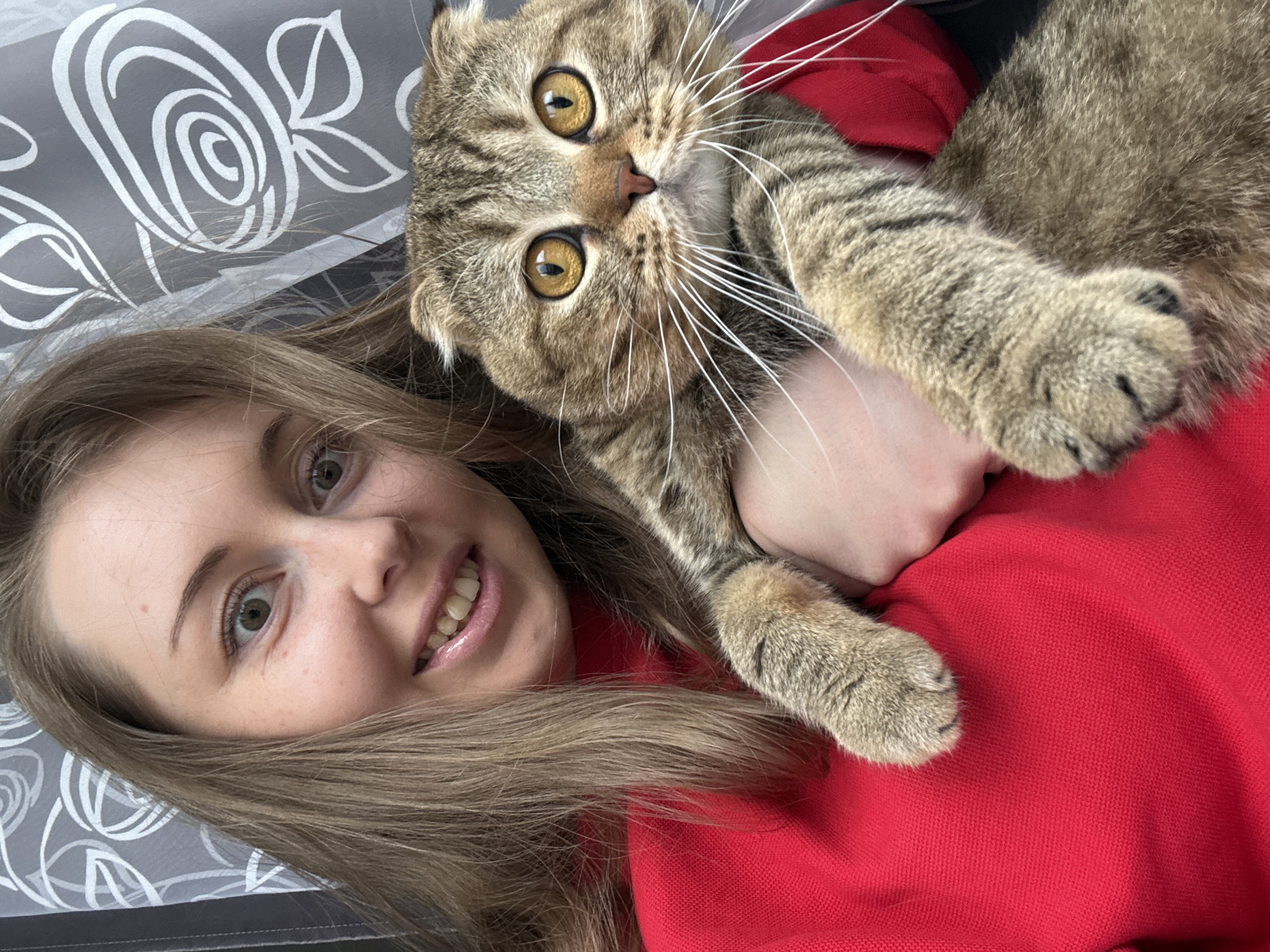 A selfie-style photograph shows a fair-skinned young woman lying on a gray and white patterned surface, possibly a bed, wearing a red shirt. A brown tabby cat with golden eyes is lying on her chest, looking directly at the camera. The woman is smiling, and her hair is light brown. The location is Unknown, Unknown.