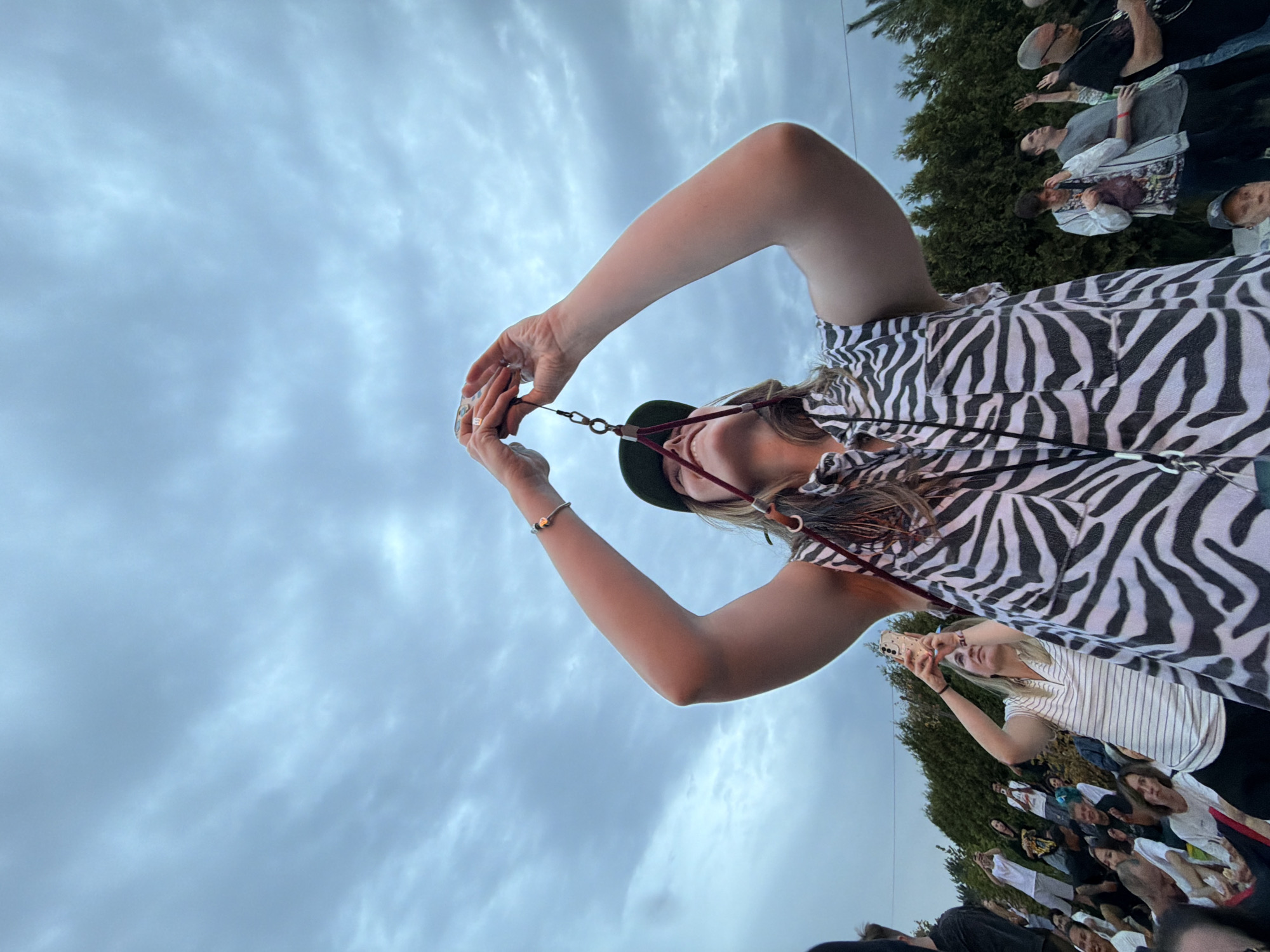 In Pervomayskiye Prudy, Kazakhstan, a low-angle shot captures a group of people outdoors under a cloudy sky. In the foreground, a woman wearing a zebra-print shirt and a dark green cap is holding up her phone with both hands to take a picture. A lanyard with a metal clasp hangs from her phone. To her left, another woman in a striped polo shirt is also holding a phone up. In the background, a crowd of people are seated or standing, with a line of trees visible behind them. Some individuals in the crowd have their hands raised. The lighting suggests it is daytime, possibly late afternoon or early evening.