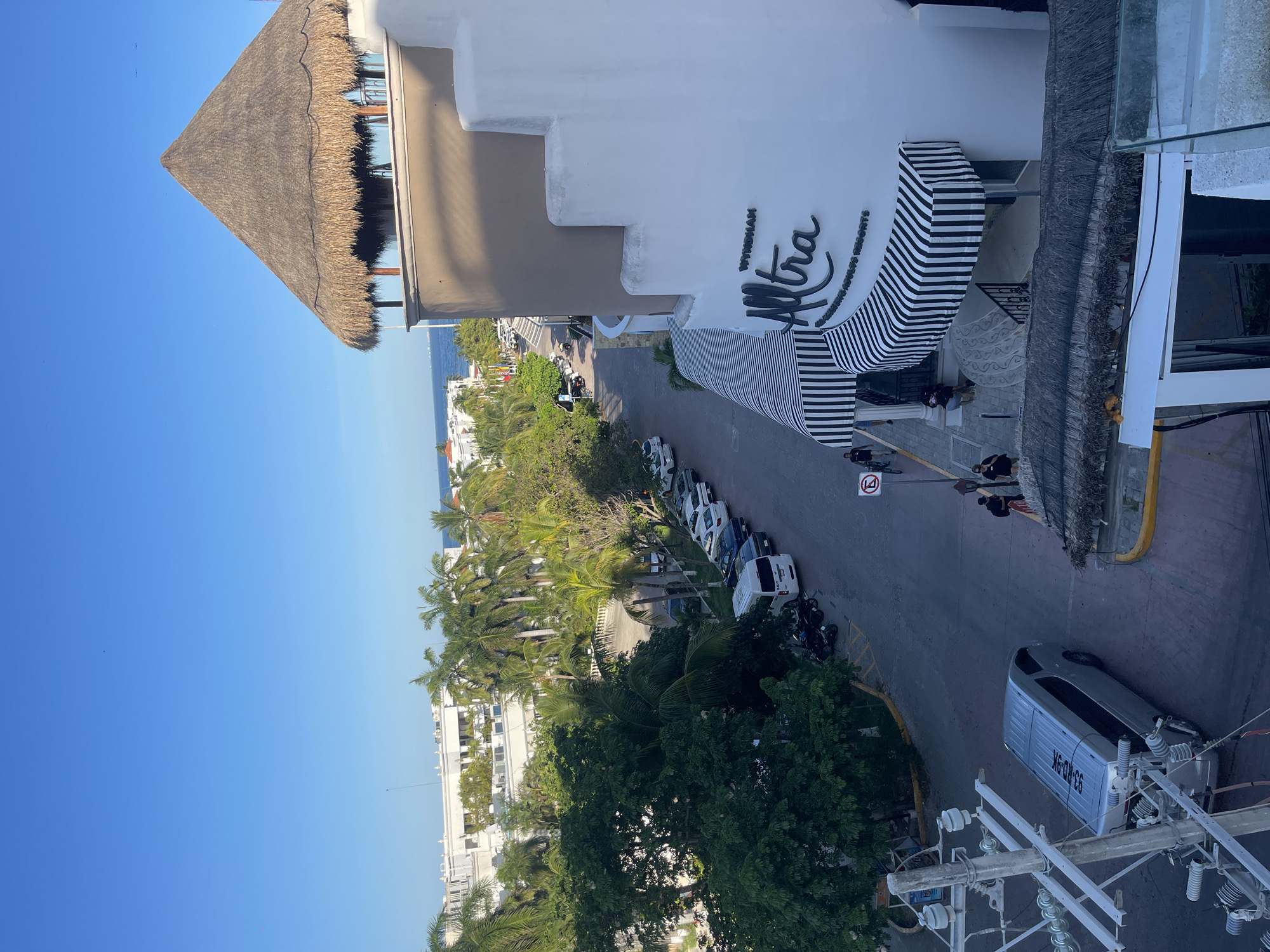 The image shows a street scene in Playa del Carmen, Mexico, with the Wyndham Alltra resort building visible on the right. The building features a white facade with a striped awning and the resort name displayed. Palm trees and lush greenery line the street, with several parked cars visible. In the distance, the ocean can be seen. Power lines and a vehicle are present in the foreground. People are walking in front of the resort. A speed limit sign is visible on the right side of the street. A building with a thatched roof is also visible on the left.