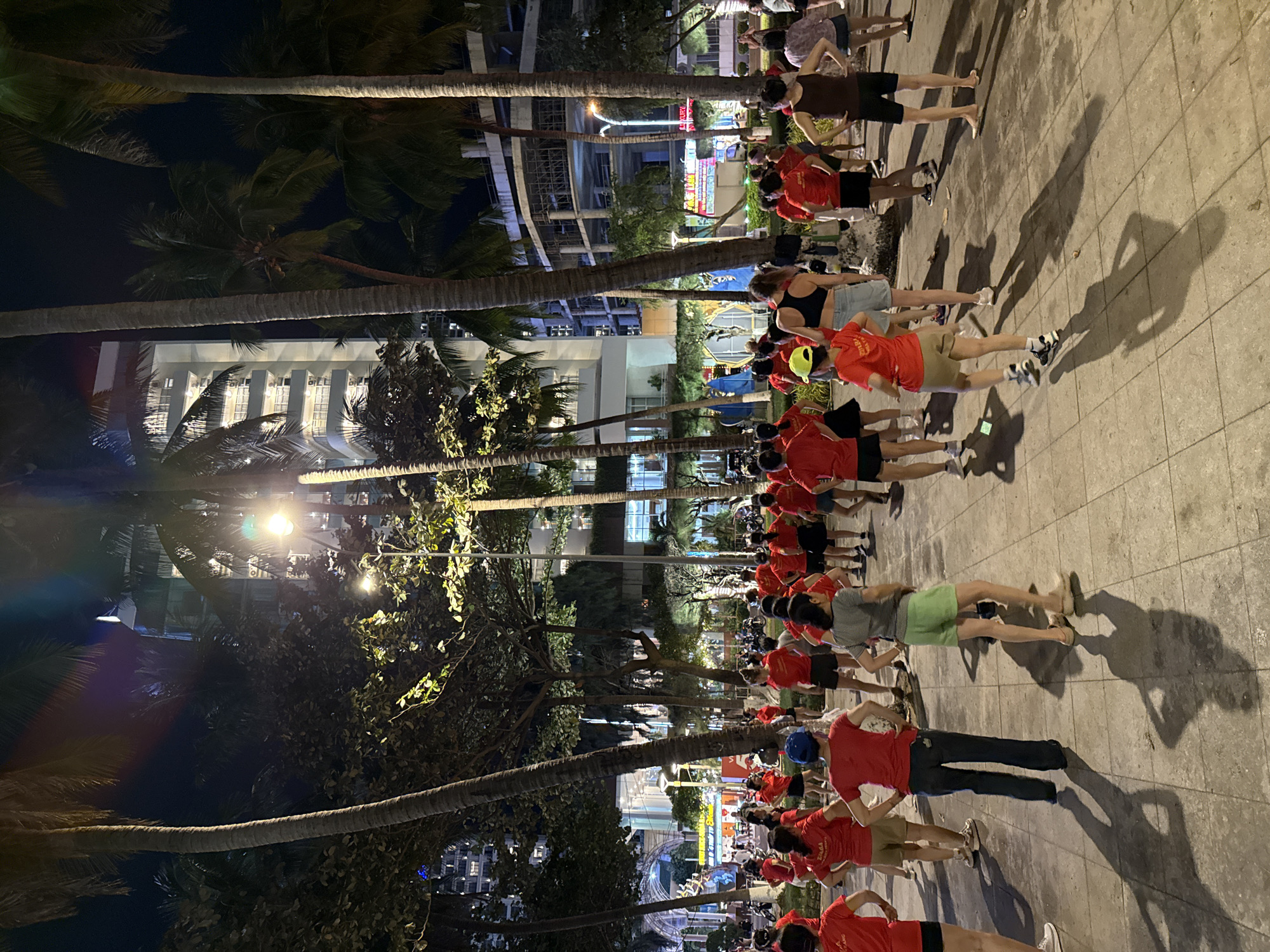 In Nha Trang, Vietnam, at night, a group of people are gathered in an outdoor area, possibly a park or plaza, near palm trees and a multi-story building under construction. The majority of the individuals are wearing red tops and dark bottoms, suggesting they are part of a coordinated activity or event. Many are engaged in what appears to be a dance or exercise routine, with some individuals in the foreground in distinct poses, hands on hips, and others facing away from the camera. The ground is paved with square tiles, and the scene is illuminated by artificial lighting, casting long shadows. In the background, there are more trees, buildings, and what looks like a street with some parked motorcycles. The overall atmosphere is one of a community gathering engaged in physical activity.