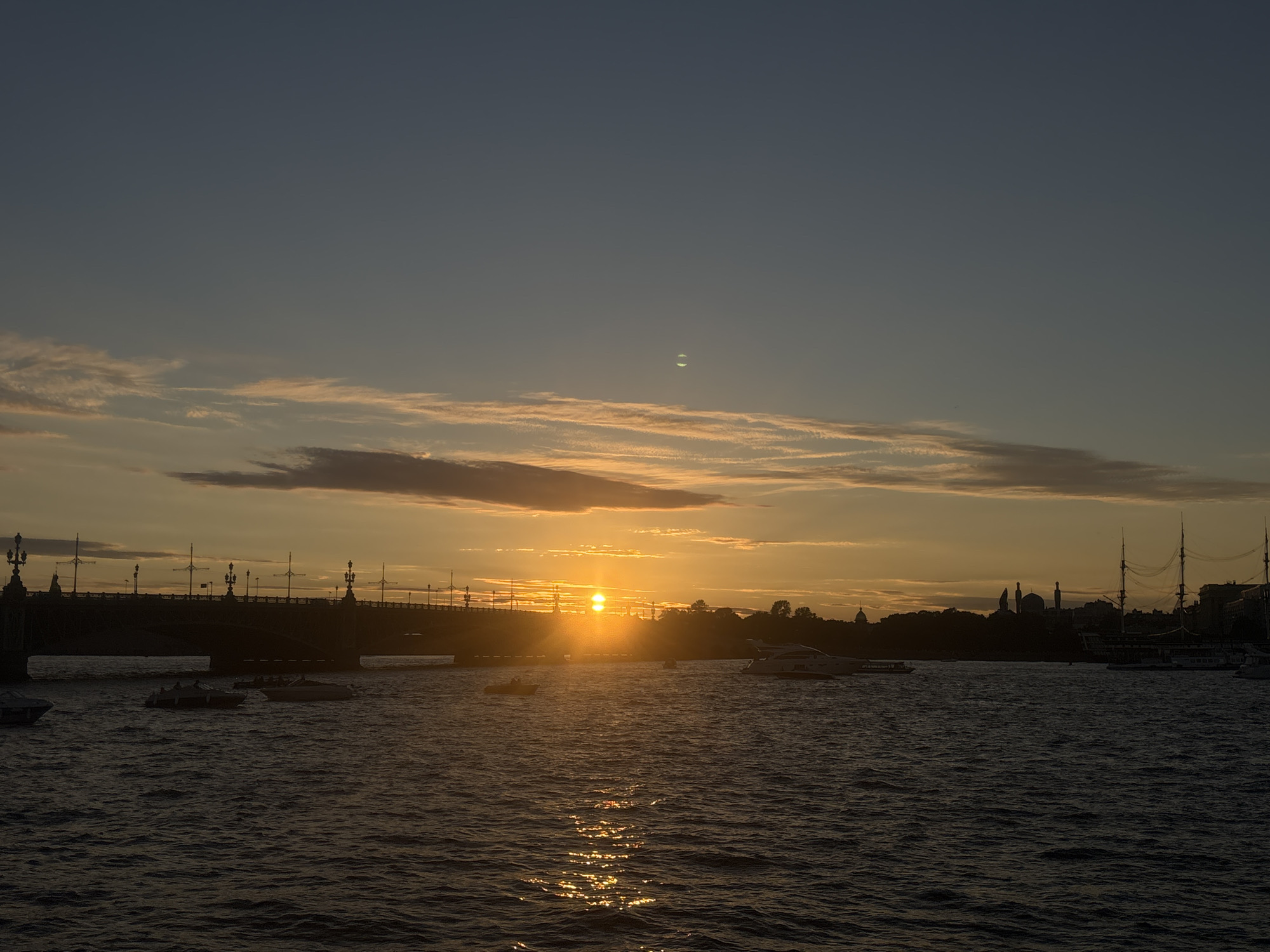 The image captures a sunset over the Neva River in Saint Petersburg, Russia, with a bridge and various boats silhouetted against the vibrant sky. The sun is low on the horizon, casting a golden glow on the water and clouds. The bridge spans across the river, with ornate lampposts visible along its length. Several boats of varying sizes are scattered on the water, some appearing to be in motion, leaving small wakes. In the distance, the city skyline is visible, with buildings and the masts of a large ship adding to the silhouette. The sky is a gradient of deep blue at the top, transitioning to orange and yellow hues near the horizon, with scattered clouds. There are no discernible people or specific actions depicted. The overall scene is serene and picturesque, characteristic of a summer evening in the city.
