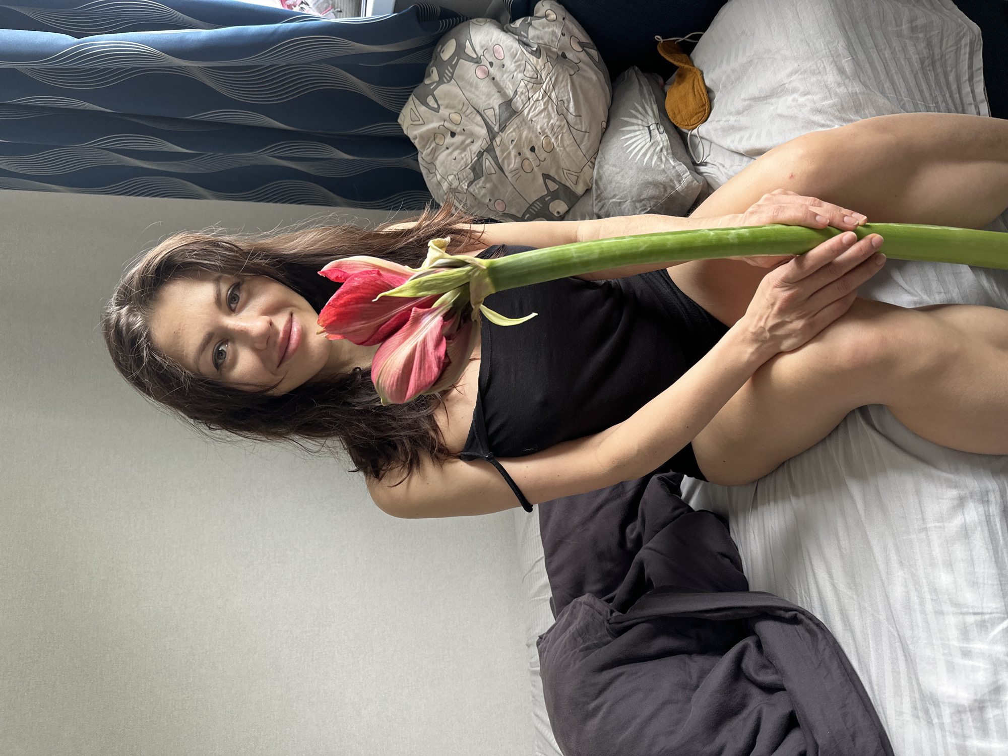 A young woman is depicted lying on a bed indoors, appearing to be in a bedroom setting. Her head is positioned on the left side of the frame, angled towards the viewer. She has long, dark brown hair and is smiling subtly, looking directly at the camera. She is wearing a black, spaghetti-strap top or bodysuit.
She is holding a large, red and pink flower with a prominent thick green stem. Her hands are clasped around the stem, which extends across her body. Her bare legs are visible, suggesting she is wearing shorts or underwear.
The setting is a bed covered with a striped white sheet and a dark grey or black blanket folded at the foot of the bed. Several pillows are visible; one is light grey with a pattern of white cats, and another is plain light grey. An orange sleep mask is resting on one of the pillows.
In the background, a window is visible on the upper left, partially covered by blue curtains with a white wavy pattern. The wall to the left of the bed is plain light grey or white. The overall impression is a relaxed, casual indoor scene during what appears to be daytime, given the natural light filtering in from the window. No visible text is present in the image.