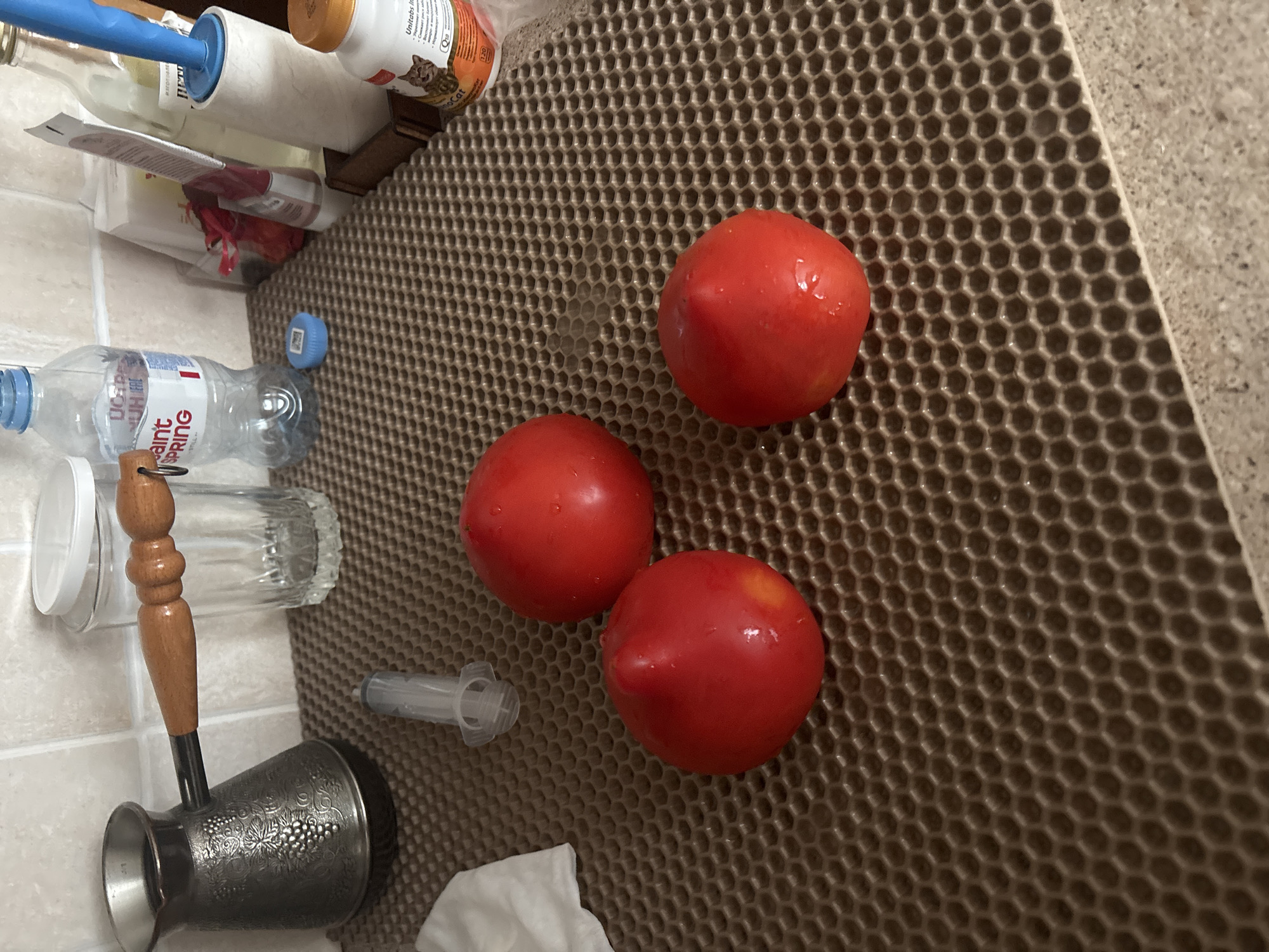The image depicts a kitchen counter with several objects. In the foreground, three red tomatoes are arranged on a brown honeycomb-patterned mat. A plastic syringe is visible between the tomatoes and a cezve with a wooden handle. In the background, a clear glass with a white lid, a plastic water bottle, and other items are present. The overall setting suggests a domestic environment, possibly a kitchen. The location is unknown and there are no people present in the image.