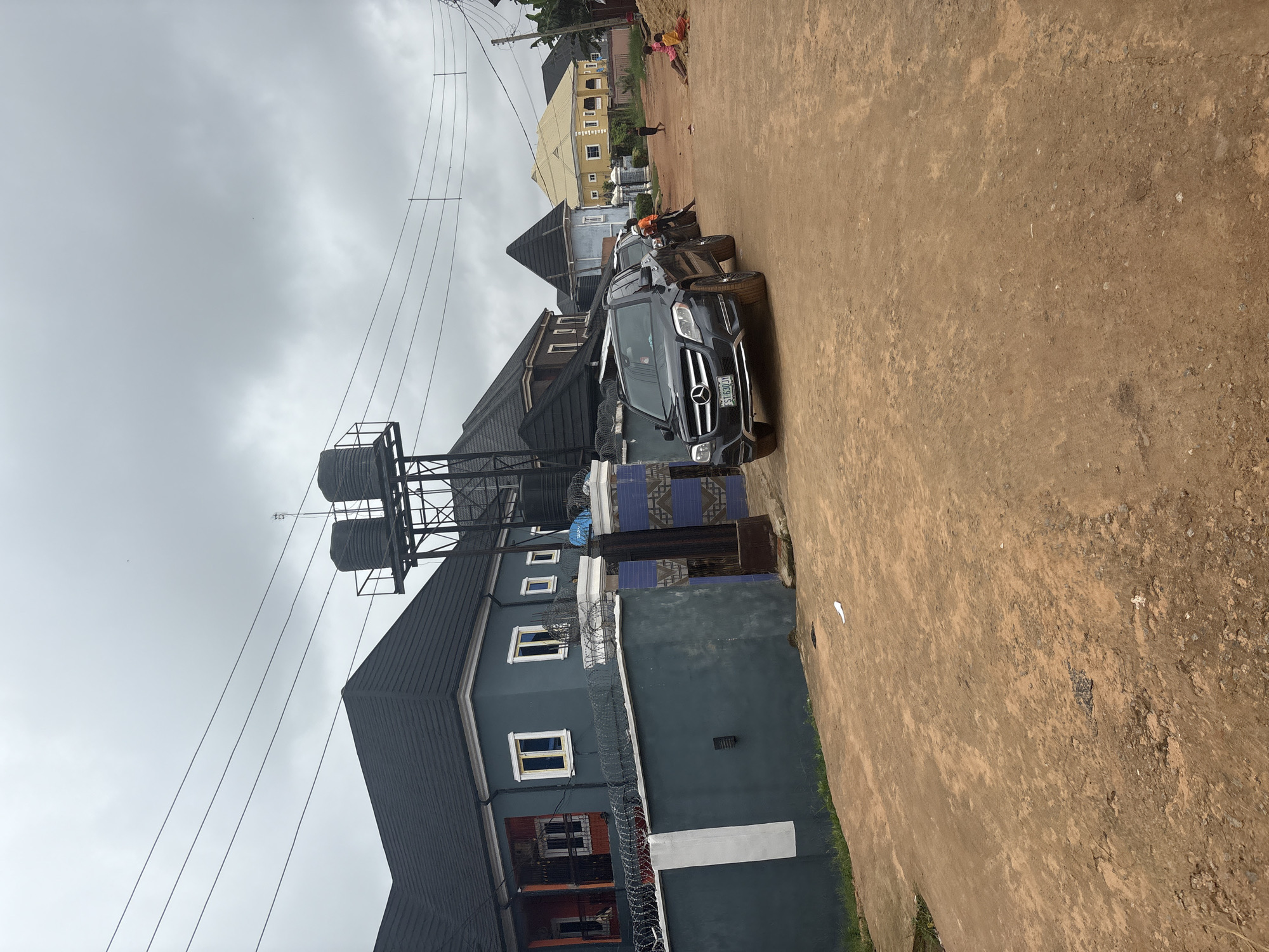 In the city of Obosi, Nigeria, on a cloudy day, a residential street is depicted. The foreground features a dirt road with tire tracks visible. To the left, a blue house with a black tiled roof is bordered by a grey wall topped with barbed wire. A dark grey Mercedes-Benz SUV is parked on the road in front of the house, with its license plate visible. Behind the SUV, other vehicles and houses are present. To the right, a group of children are sitting on a dirt pile and on the ground. Power lines run across the sky.