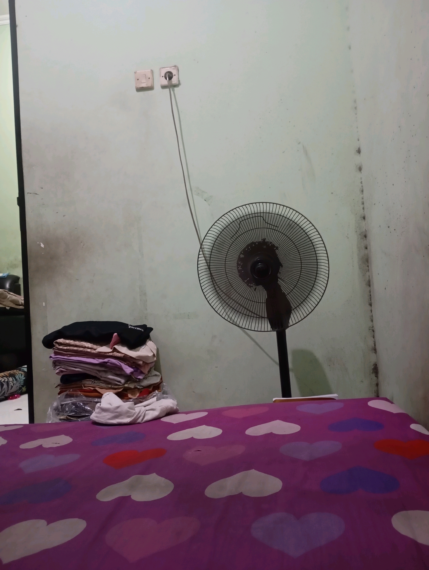 A static shot captures a corner of a room in Babelan, Indonesia. The wall, painted a faded mint green, shows signs of wear and tear, with dark smudges and water stains visible. Two electrical outlets are mounted on the wall; one is a simple switch, while the other has a plug inserted into it, connected to a black cord that trails down to a standing fan. The fan, a dark color, is positioned to the right of the frame, its circular grille and blades clearly visible.
To the left of the fan, a stack of folded clothes sits on a surface that is partially obscured. The clothes are a mix of colors, primarily muted tones of pink, purple, and white, topped with a black garment. Below the clothes, a clear plastic bag is visible, containing more items.
The foreground of the image is dominated by a bed covered with a vibrant purple sheet adorned with a repeating pattern of multi-colored hearts in shades of pink, purple, red, and white. A light-colored folded garment rests on the edge of the bed. The overall impression is one of a domestic, lived-in space.