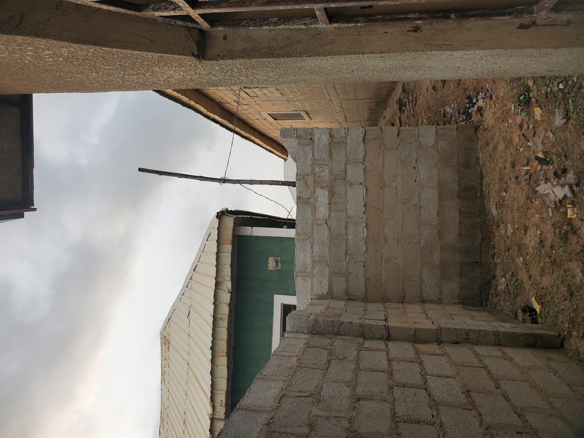 This media file shows an outdoor scene, likely in Suleja, Nigeria. The image is oriented vertically, with the ground appearing at the top and the sky at the bottom.
The foreground is dominated by the construction of a wall made of concrete blocks. To the left, a portion of a building with a corrugated metal roof is visible. This building has a green facade and a small window or fixture. In the background, a section of another building with a tiled roof and a small rectangular window is seen. Above this, a light pole extends upwards against a cloudy sky.
The ground next to the block wall is unpaved and appears to be littered with various debris, including plastic bags and other refuse. The overall impression is of an ongoing construction site or an area that is not well-maintained.
The sky is overcast, suggesting it might be a cloudy day or late afternoon. There are no people or animals visible in the frame, and no specific activity is taking place beyond the implied construction. The scene conveys a sense of incompleteness and the urban environment of a developing area. No text is visible in the image.