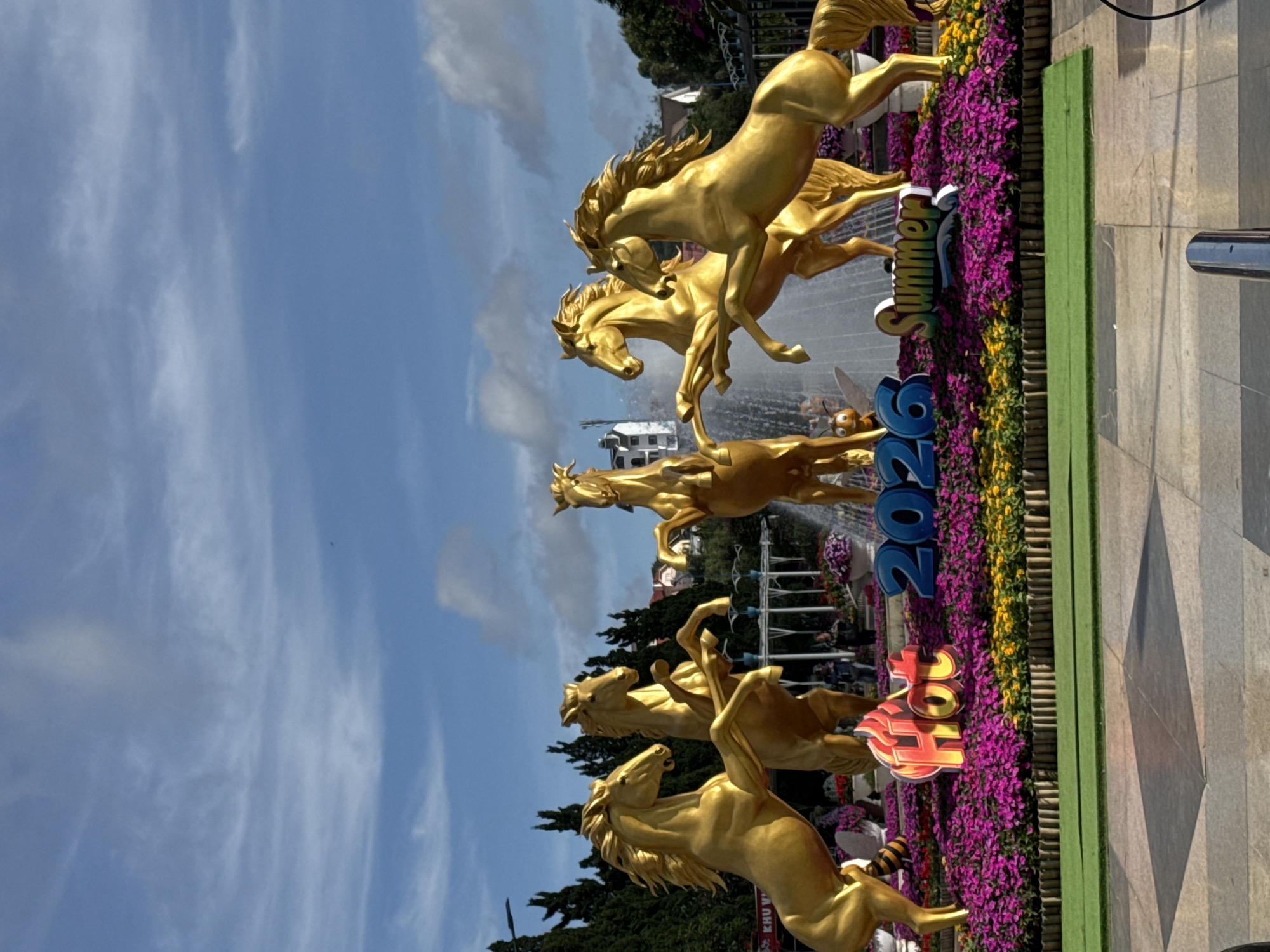 This image displays an outdoor public art installation featuring six golden horse sculptures, a central fountain, and extensive floral arrangements. Five golden horses are prominently positioned in a rearing posture around the fountain, with one additional horse partially visible behind it. The sculptures exhibit a metallic golden finish. A multi-tiered fountain is centrally located, actively spraying water. Surrounding the fountain and horses is a raised flower bed densely planted with vibrant magenta-purple flowers, bordered by a strip of yellow flowers and a low wooden fence. Within the flower bed, three distinct signs are visible: "Hot" in flame-stylized red and orange letters on the left, "2026" in blue block numbers centrally, and "Summer" in stylized green and yellow letters on the right. A small cartoon bee figure is positioned near the "Hot" sign. The foreground consists of light grey paved ground with darker square patterns, and a strip of artificial green turf. In the background, dark green coniferous trees and several white and light-colored buildings with contrasting roofs are visible under a bright blue sky with scattered white clouds. A few individuals are discernible in the midground behind the sculptures, observing the display. The scene is well-lit by natural daylight. This installation is located in Phường Xuân Hương, Đà Lạt, Vietnam.