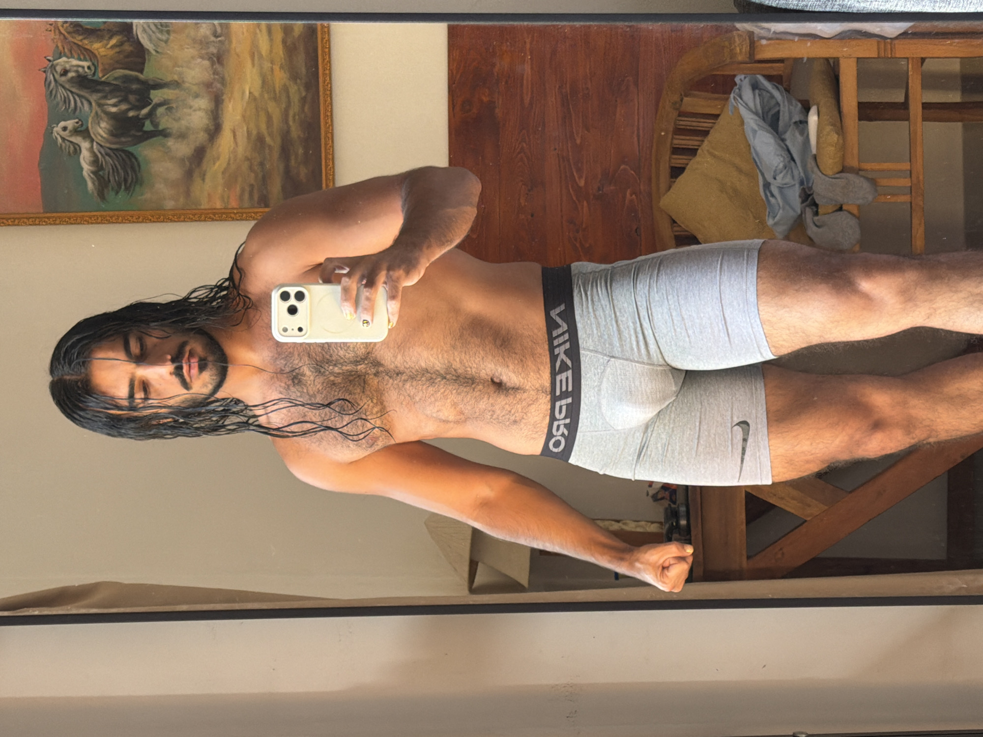 A shirtless man with long, dark, wet hair and a beard stands in front of a mirror, taking a selfie with a smartphone. He is wearing grey boxer briefs with a black waistband. His left arm is bent, with his fist clenched. In the background, a painting of horses is visible on the wall. A lamp and a wooden chair with a cushion are also present. The scene appears to be indoors, likely in a residential setting. No specific location in North Kuta, Indonesia, is identifiable from the image.
