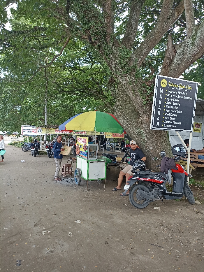 This image depicts an informal outdoor eatery, commonly known as a *warung*, in Kepung, Indonesia, under the shade of a massive, ancient tree during the daytime. The ground is unpaved, consisting of dirt and gravel.
In the foreground, a man in a blue shirt stands next to a green and white food cart, which is topped with a vibrant yellow, green, and red umbrella. He looks towards the camera with a slight smile, holding a flat, tray-like object. To his right, another man is seated on a low stool or bench, casually leaning against the tree trunk. He wears a dark t-shirt, shorts, and a baseball cap, and a black and red scooter is parked in front of him. On the far left, a woman is partially visible walking away. Several other motorcycles are parked further back, near additional simple stalls and structures.
A large black sign, numbered "354," is affixed to the tree trunk. It displays "Warung Nada Cinta" at the top and lists a menu under "MAKANAN" (Food). The listed dishes include "Rica-rica Menthok," "Rica-rica Ayam Kampung," "Ayam Bakar," "Ayam Goreng," "Nasi Wader," "Nasi Pecel Telur," "Nasi Kumlag," "Nasi Liwet," "Sambal Pindang," and "Sambal Teri." In the background, a white sign is partially visible, offering "GRATIS MINUMAN" (Free Drinks) and "FREE WIFI." The overall scene suggests a casual and popular local gathering spot for food and relaxation.