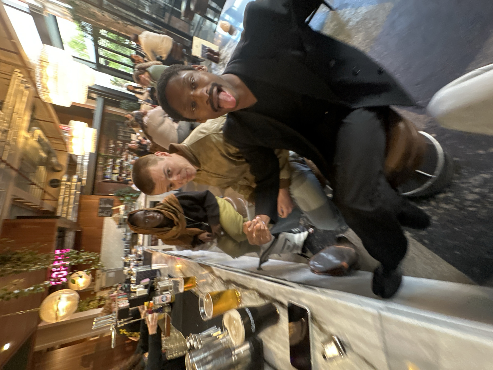 The image shows a group of four people, presumably in a bar or restaurant, situated in the city of London, United Kingdom.
From foreground to background, the scene includes:
1. **Bar Area:** A counter with glassware, including pint glasses and what appears to be a cocktail shaker. There is also a dark smartphone with a screen facing away from the camera. Behind this counter, there is a person in black attire serving drinks.
2. **People:** Four people are prominent in the foreground. The person closest to the camera is wearing a black blazer and appears to have his tongue out. Behind him, another person is wearing a tan jacket. Behind these two there is a person with a brown hooded sweatshirt with a black shirt. A forth person is wearing a light tan jacket.
3. **Restaurant Interior:** Background details reveal tables and seating with other patrons. Lighting fixtures, including chandeliers or hanging lamps, are present in the room. The architecture appears modern and stylish.
4. **Location:** The urban setting is confirmed by the style of the interior, which aligns with establishments often found in London.