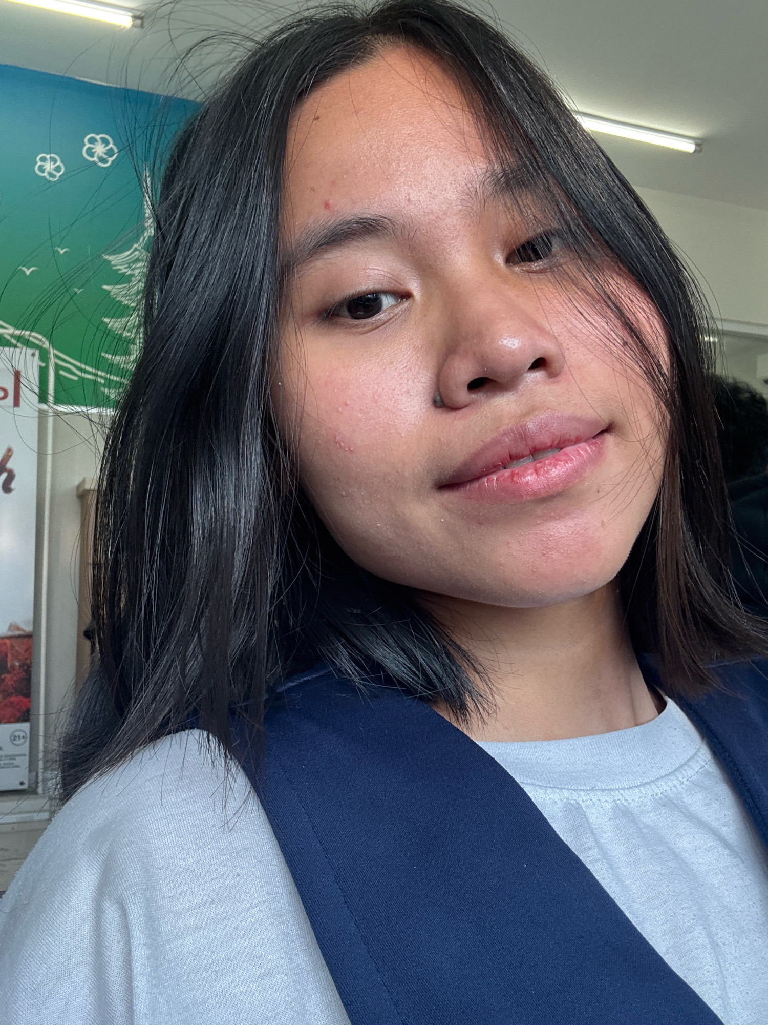 The image shows a close-up selfie of a young woman with medium-length dark hair. She is wearing a light-colored t-shirt and a dark blue vest or jacket. The background appears to be indoors, with a brightly lit ceiling and a wall decorated with a green mural featuring white floral designs and birds. The woman's face is slightly tilted, and she is looking towards the camera with a subtle smile. There are some minor blemishes visible on her skin. The overall impression is that of a casual, personal photograph taken in an urban indoor setting. The specific location in East Jakarta, Indonesia, cannot be determined from the visual information provided.