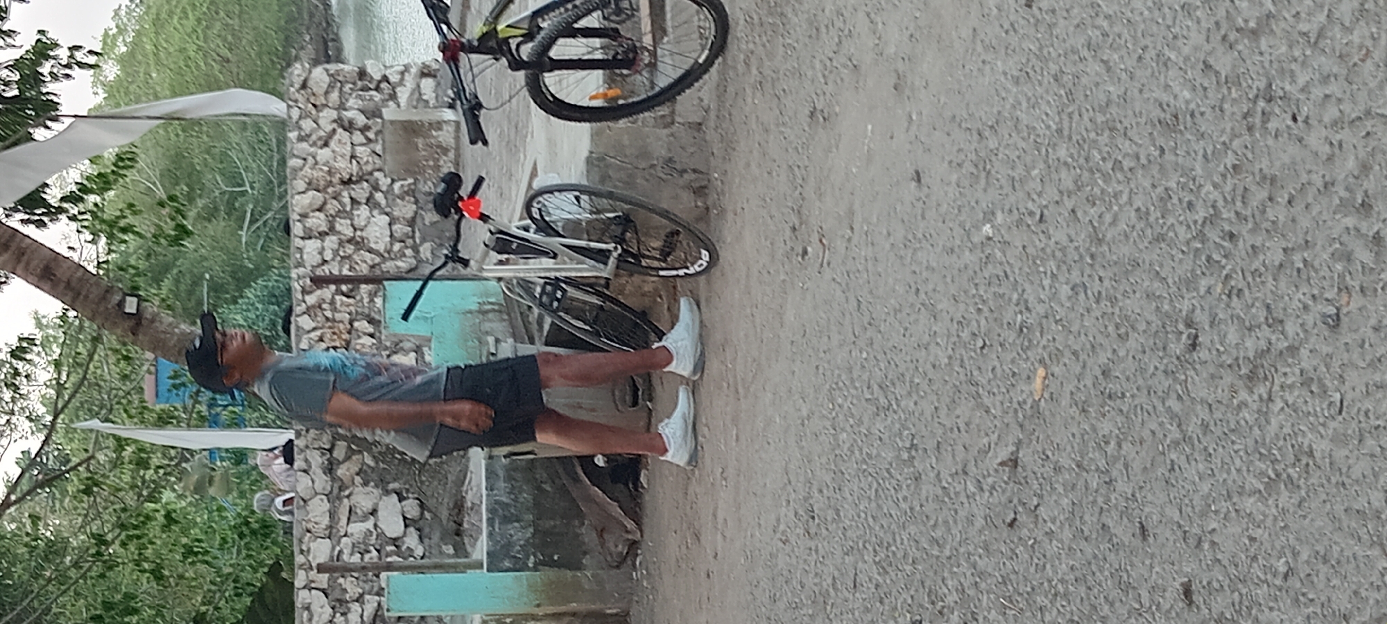 The image depicts a man standing next to two bicycles in what appears to be an outdoor area with a stone wall and foliage. The man is wearing a dark cap, glasses, a dark t-shirt, and dark shorts with white athletic shoes. He is facing to the right of the frame, looking slightly upwards. In the background, a stone wall is visible, behind which is lush green vegetation and trees. A white flag or banner hangs from a tree. One bicycle is silver and black with a red reflector on the handlebars, and the other is black and yellow. The ground is covered in gravel and dirt. The setting suggests a casual outdoor environment, possibly near a beach or a park, given the presence of water in the distance and the tropical vegetation. The overall atmosphere is relaxed and informal. The location is identified as Kalipucang, Indonesia.
