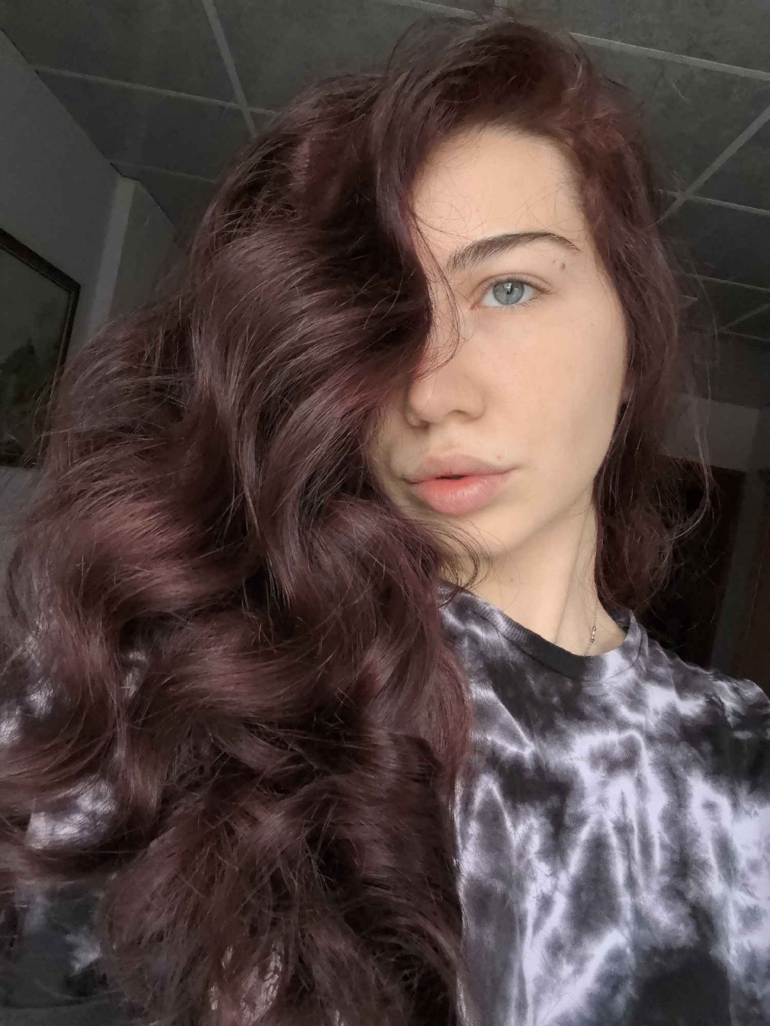 A close-up indoor portrait depicts a female individual in Stara Zagora, Bulgaria, from the upper chest upwards. She possesses long, wavy, dark red hair, parted on the right, with a significant portion draped over the left side of her face, partially obscuring her left eye and temple. Her visible right eye is light blue, and her eyebrows are defined. Her skin complexion is fair, and her lips are full with a natural, light pink hue. She is wearing a short-sleeved t-shirt featuring a grey and white tie-dye pattern. A delicate silver-colored chain necklace is visible around her neck. The background consists of a light-colored wall on the right and a darker wall on the left, where a framed object, possibly a picture or mirror, is partially visible. The ceiling, featuring light-colored panels with visible seams, is present in the upper portion of the frame. The lighting source appears to be from the front or slightly to the right of the subject, creating soft shadows. The individual is looking directly at the camera with a neutral expression.