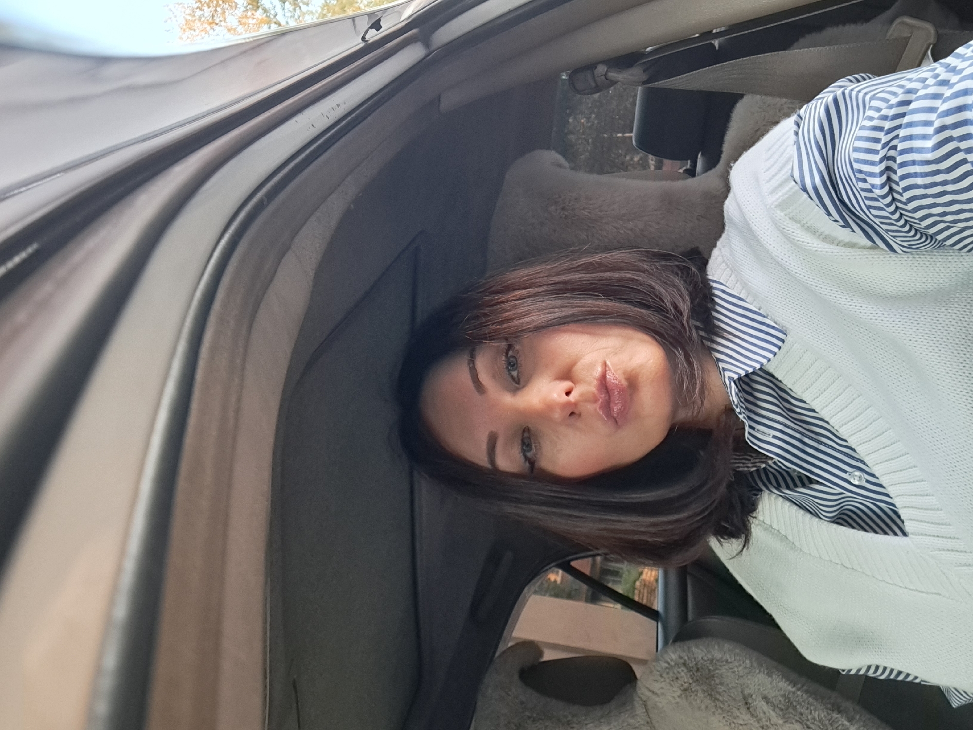 A woman is pictured inside a car. She is looking at the camera and has her lips pursed. She is wearing a blue and white striped shirt and a white sweater vest. The car appears to be parked outdoors on a sunny day. The interior of the car is visible, including the seats and the roof lining. The background shows trees and a building, suggesting an urban or suburban environment. The woman's hair is dark brown and medium length.