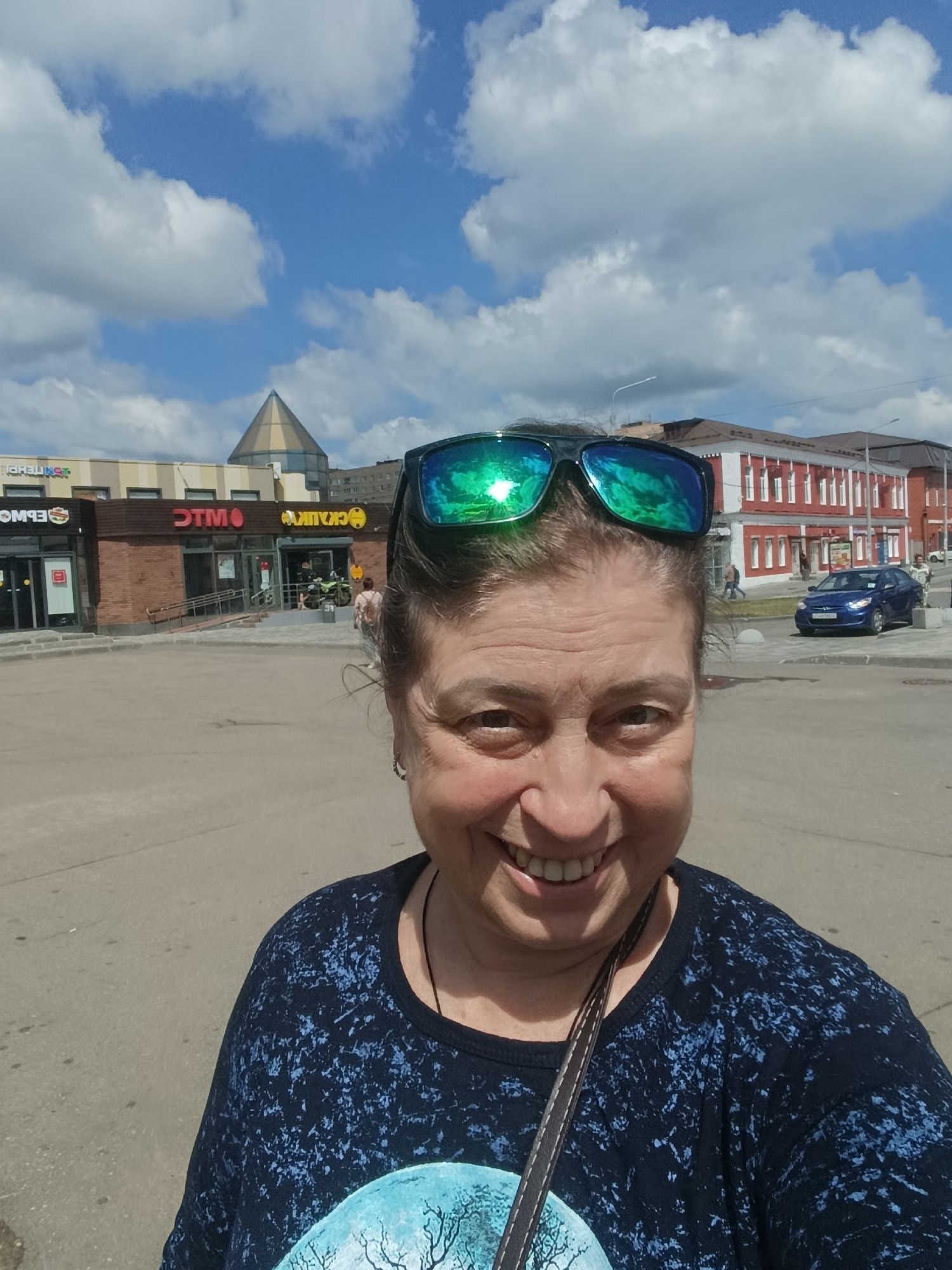 The image shows a woman taking a selfie outdoors in Orekhovo-Zuyevo, Russia. She is wearing sunglasses and a dark blue t-shirt. In the background, there are buildings with signage, a blue car, and a clear sky with clouds. The environment appears to be an urban area with a paved ground surface.