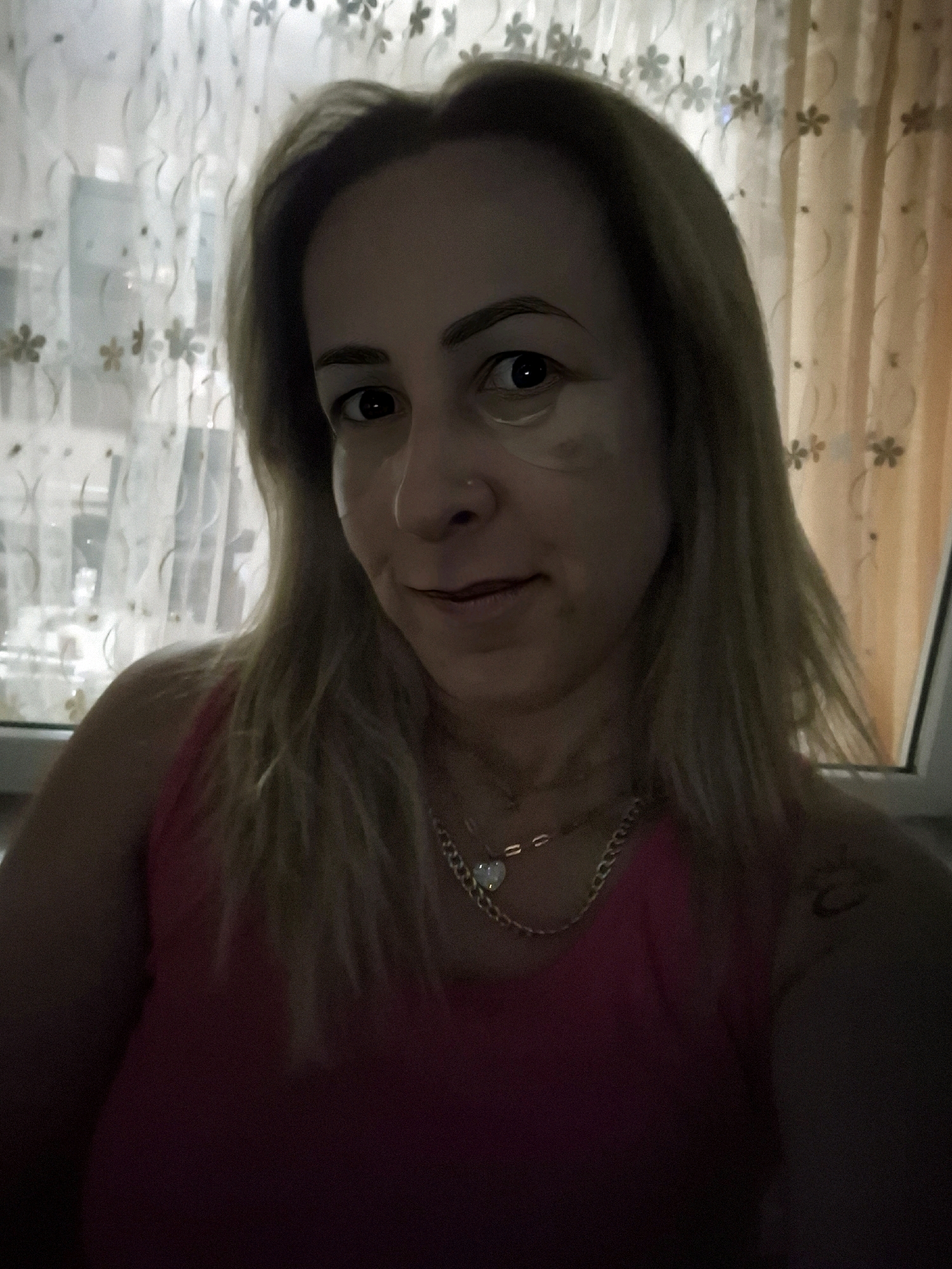 A close-up, selfie-style photo shows a woman with blonde hair and dark eyes. She is wearing a pink top and a chain necklace with a heart pendant. There are light-colored curtains with floral patterns in the background, and a window is visible to the right. The woman has a nose ring and what appear to be under-eye patches.