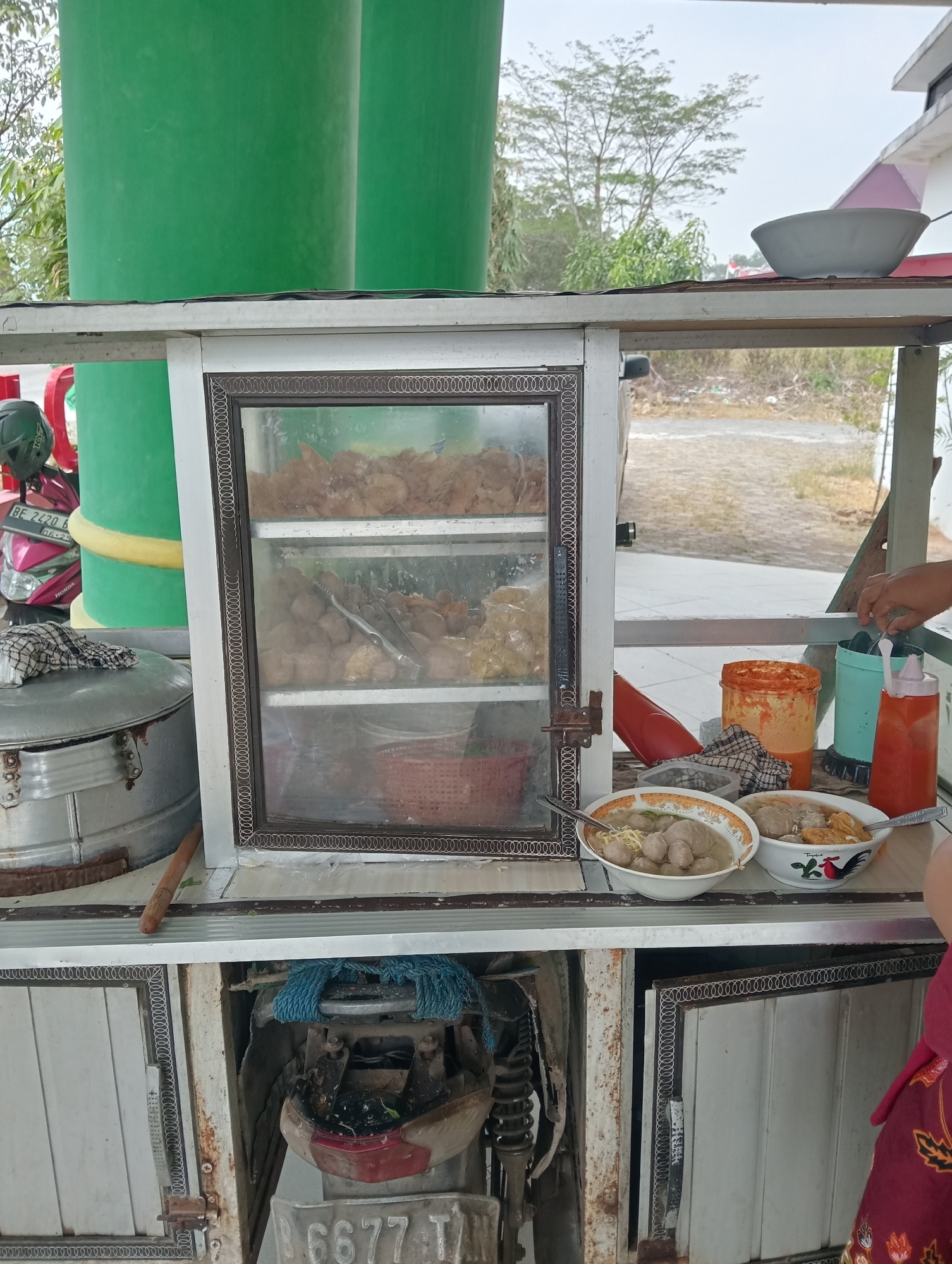 A food cart is parked outdoors, likely in a city like Sidoharjo, Indonesia. The cart features a glass display cabinet filled with various food items, including what appears to be fried chicken and meatballs. Below the display are storage compartments. On the counter of the cart, two bowls of soup, likely bakso, are visible, along with condiments such as chili sauce and a container of orange paste. A person's hand is reaching for one of the bowls. In the background, two large green pillars stand, and trees and a building suggest an urban or semi-urban setting. A motorcycle is parked to the left of the cart. The overall scene depicts a street food vendor preparing to serve customers.
