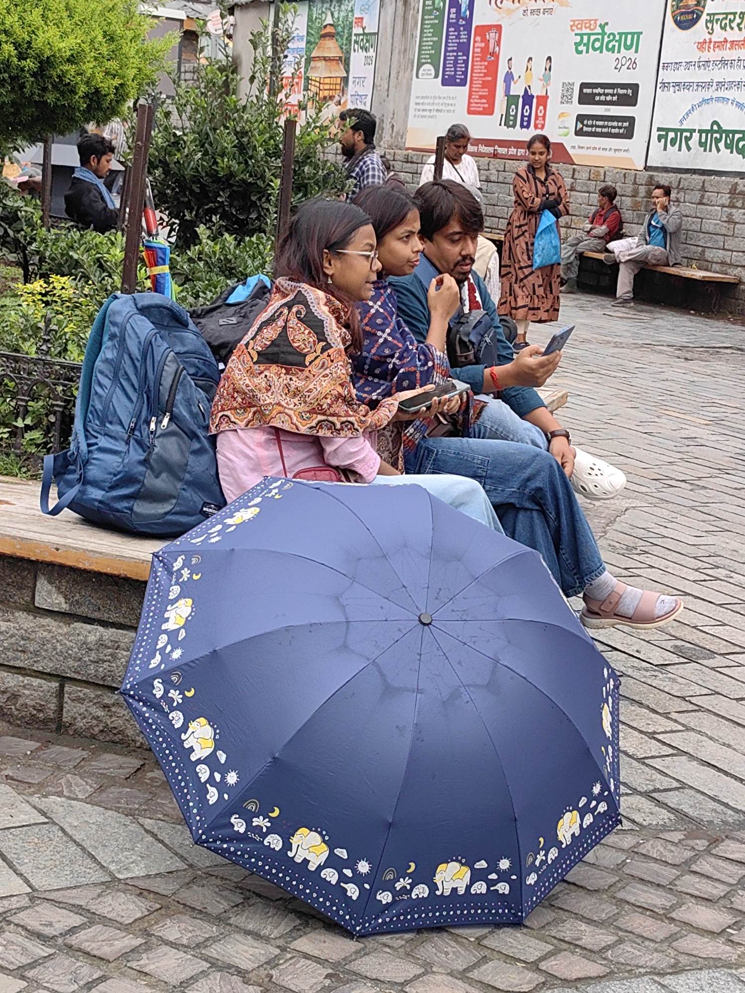 In Manali, India, a group of people are seated on a wooden bench in front of a stone wall adorned with posters. A dark blue umbrella with yellow elephant motifs rests on the ground in the foreground. Two young women and a man are seated together, with the man holding a mobile phone and interacting with it. A blue backpack is placed next to the bench. In the background, several other individuals are visible, some standing and some seated, with lush green foliage and buildings in the distance. The scene depicts a casual public gathering in an outdoor setting, possibly a park or waiting area.